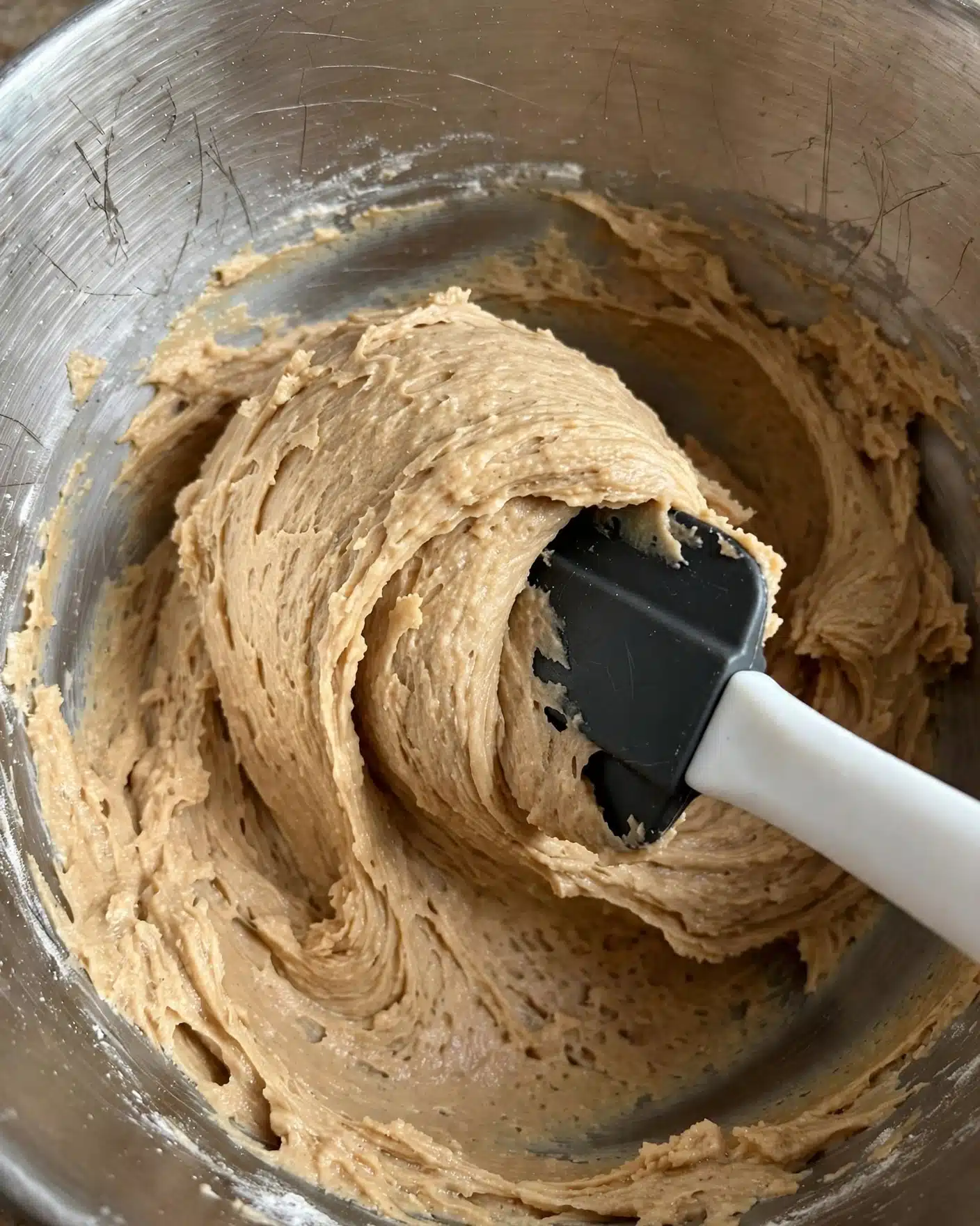 Cookie Dough Frosting Recipe 4 A close-up image shows light brown creamy dough inside a metal mixing bowl. The dough has a smooth but slightly textured look with some visible ridges and folds, pushed and spread around the sides of the bowl. A black spatula with a white handle stirs the dough, lifting some of it in a wave shape. The mixing bowl's inside surface has a slightly scratched metal look. The photo is taken with an iphone --ar 4:5 --v 7