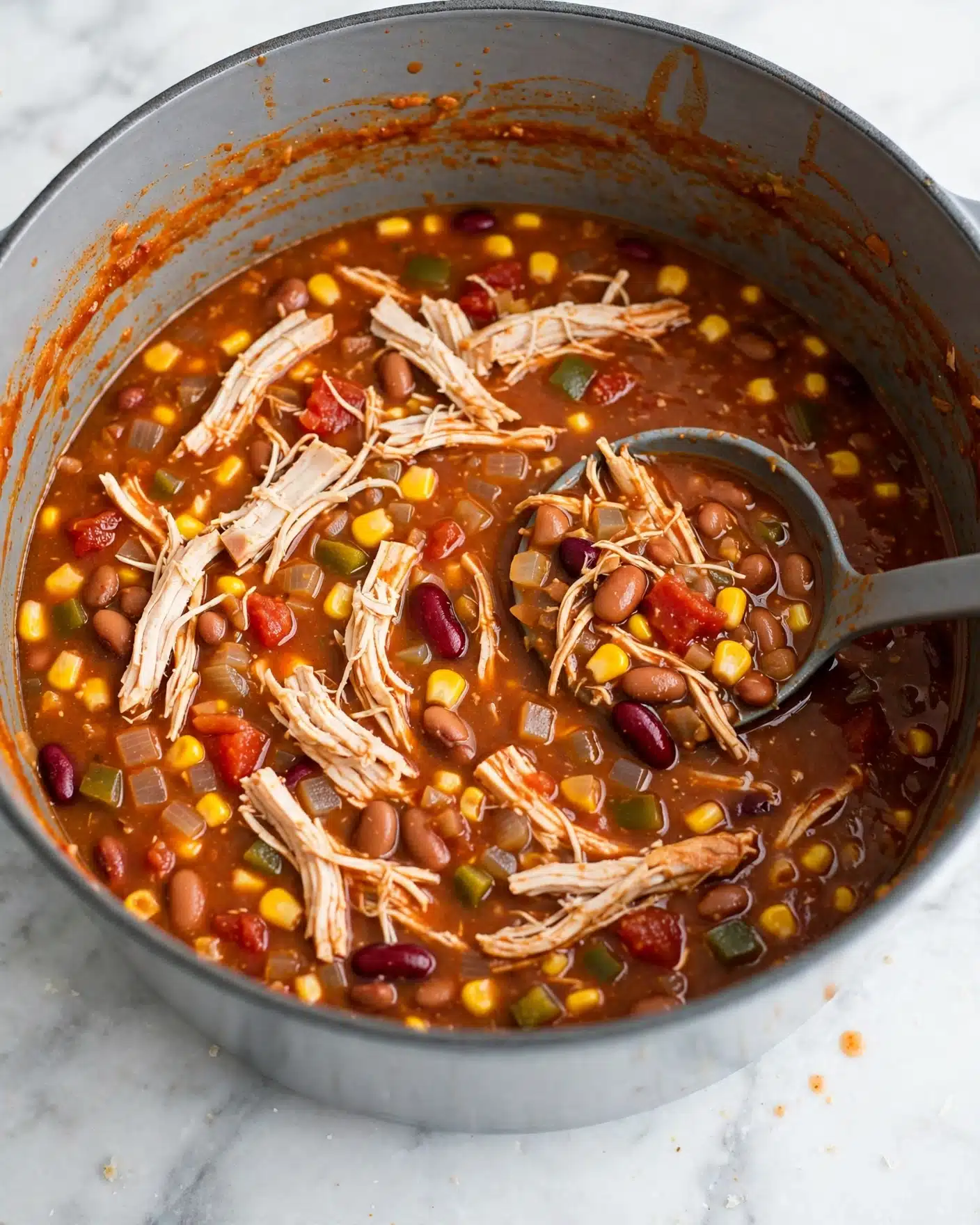 Inside a large round gray pot on a white marbled surface, there is a thick soup with several visible layers including shredded light-colored chicken spread across the top and mixed throughout, bright yellow corn kernels scattered evenly, small chunks of red tomatoes, and beans in shades of brown and dark red. The broth is rich and reddish-brown with small bits of cooked onions and green peppers visible. A gray ladle rests inside the pot, partially submerged in the soup, holding a mix of shredded chicken and vegetables. The inner sides of the pot have an orange residue from the soup, showing a thick, hearty texture. Photo taken with an iphone --ar 4:5 --v 7