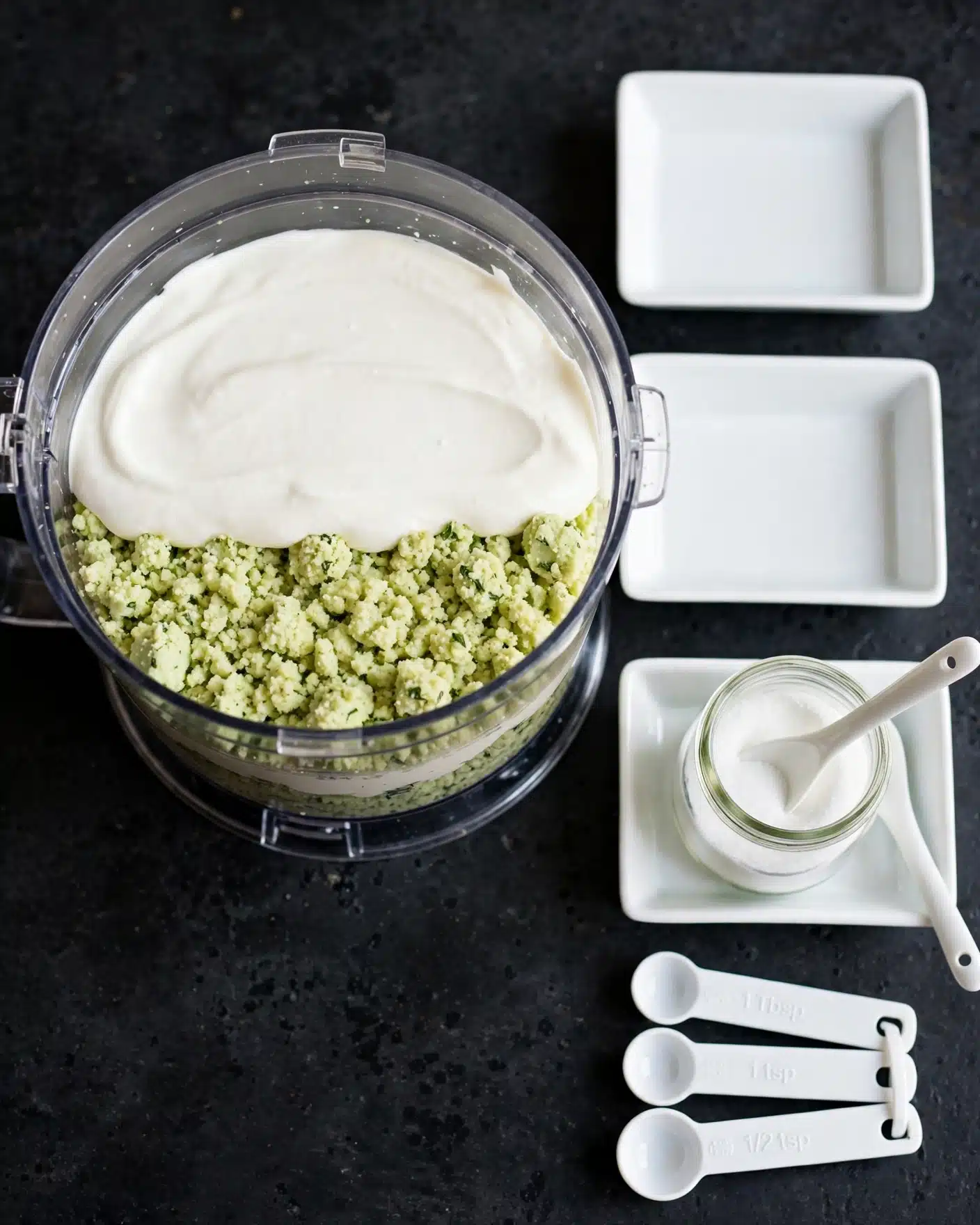 The image shows a clear food processor bowl filled with two main layers of ingredients: a crumbly light green layer of cheese on the bottom and a smooth white creamy layer of sour cream or yogurt on top. The bowl sits on a dark surface, beside three empty white rectangular dishes and white measuring spoons. A glass jar with white contents and a spoon rest in one of the dishes. The whole scene is lit from above. Photo taken with an iphone --ar 4:5 --v 7
