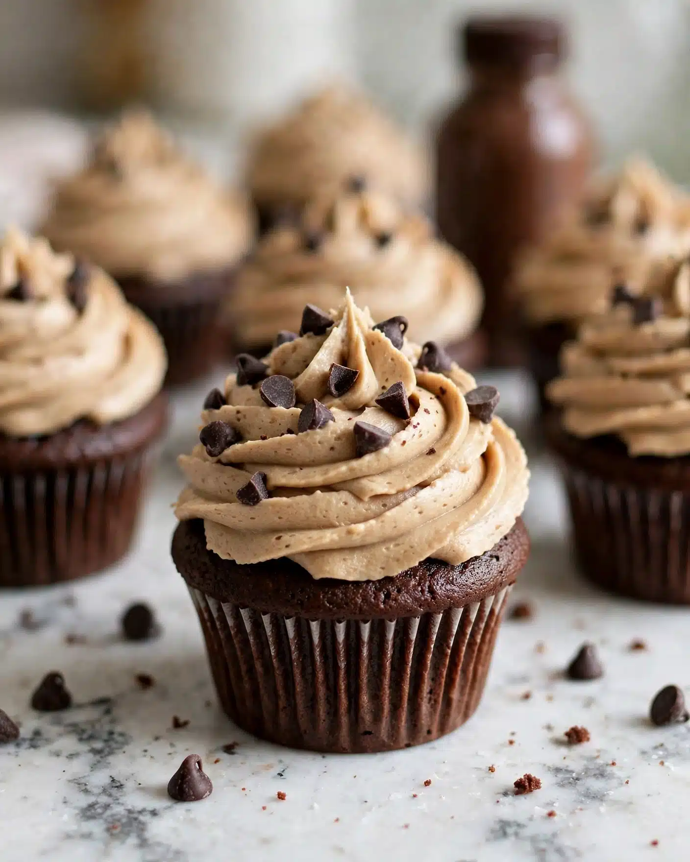 Cookie Dough Frosting Recipe 5 A close-up view of several chocolate cupcakes on a white marbled surface showing one cupcake in the front center. Each cupcake has a dark brown chocolate base with a slightly ridged texture and a thick swirl of light brown frosting on top, decorated with small dark chocolate chips scattered on and around the frosting. The background is softly blurred with more cupcakes and a brown bottle, emphasizing the central cupcake in the sharp focus. Photo taken with an iphone --ar 4:5 --v 7