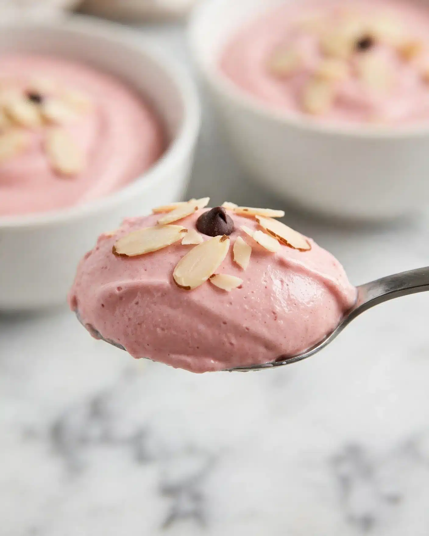 This image shows a close-up of a spoon holding a scoop of smooth, pink mousse or cream topped with small pieces of sliced almonds and one dark chocolate chip. The mousse has a creamy, soft texture with a slight shine. In the blurred background, there are white bowls filled with more of the same pink mousse, also sprinkled with almond slices. The setting is on a white marbled surface with a clean and simple look. Photo taken with an iphone --ar 4:5 --v 7