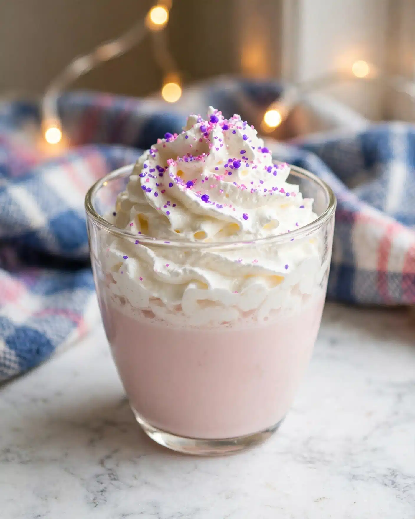 A clear glass cup holds a two-layer drink with a smooth pale pink bottom layer and a thick white whipped cream top layer. The whipped cream is generously swirled and sprinkled with tiny bright purple and pink sugar crystals, adding texture and color contrast. The glass sits on a white marbled surface with a soft plaid cloth in the background with blue, white, and pink tones, and small glowing string lights add a warm ambiance. photo taken with an iphone --ar 4:5 --v 7