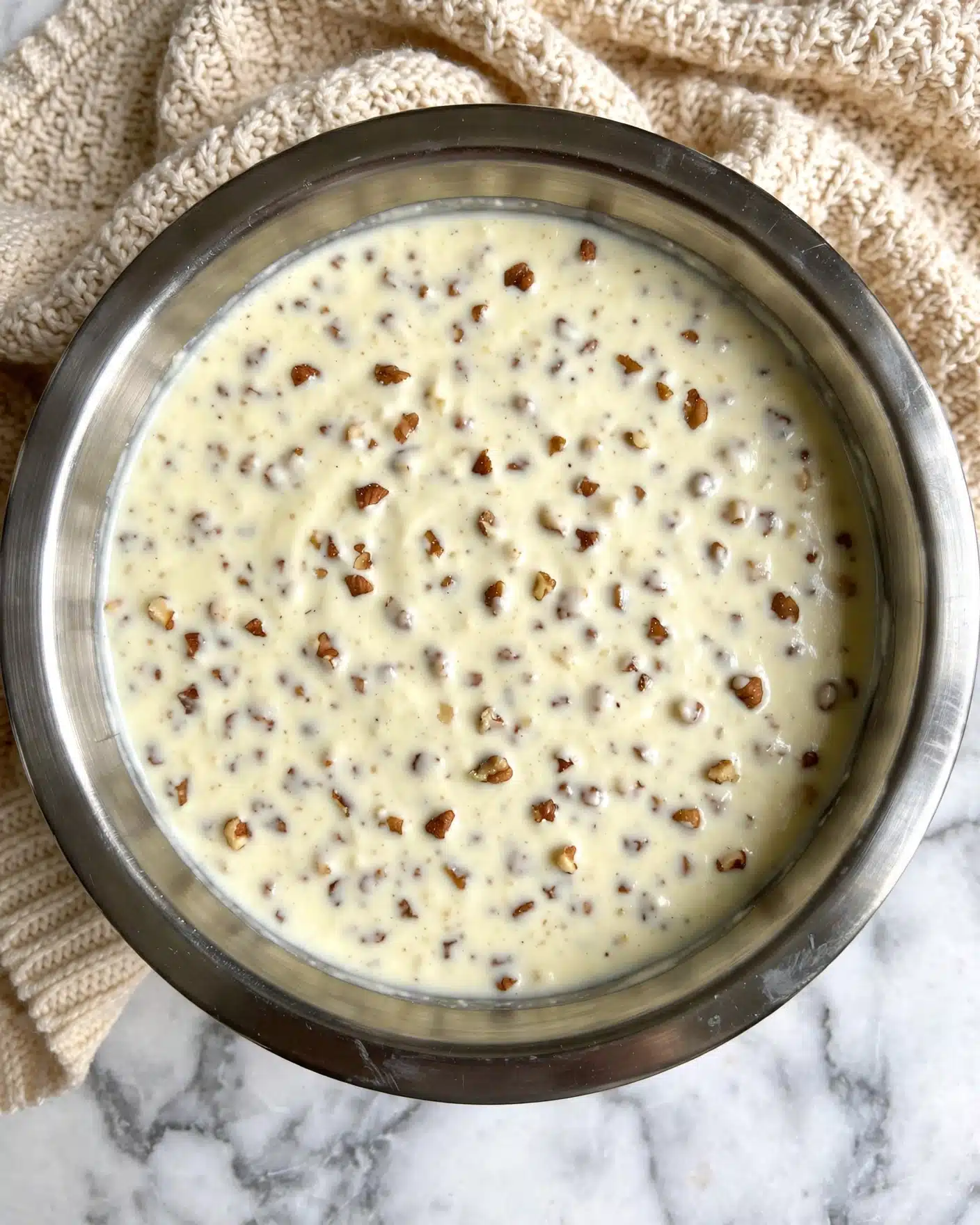 White Texas Sheet Cake Recipe 4 A close-up view of a steel bowl filled with a creamy mixture that contains many small brown nut pieces spread evenly throughout. The mixture is light yellowish-white with a smooth, thick texture. The bowl is placed on a soft, beige knitted cloth, lying on a white marbled surface. photo taken with an iphone --ar 4:5 --v 7