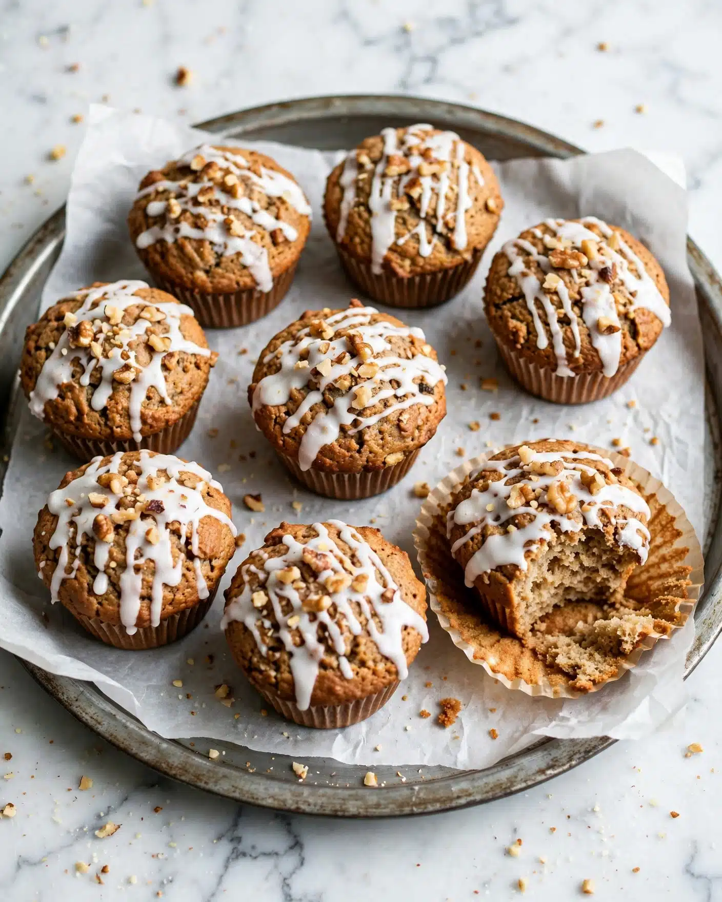 Pumpkin Carrot Cake Muffins with Spiced Vanilla Glaze Recipe 5 The image shows a group of seven muffins with a brown, textured base and a drizzle of white icing on top. The icing covers the top surface unevenly, with some areas thicker and some showing the muffin underneath. Small pieces of chopped nuts are scattered on the icing, adding texture and contrast. One muffin is peeled halfway out of its brown paper liner, revealing the detailed crumb inside, and it has a bite taken out of it. The muffins rest on white parchment paper inside a round metal tray, placed on a white marbled surface. Some crumbs are scattered around for a natural look. photo taken with an iphone --ar 4:5 --v 7