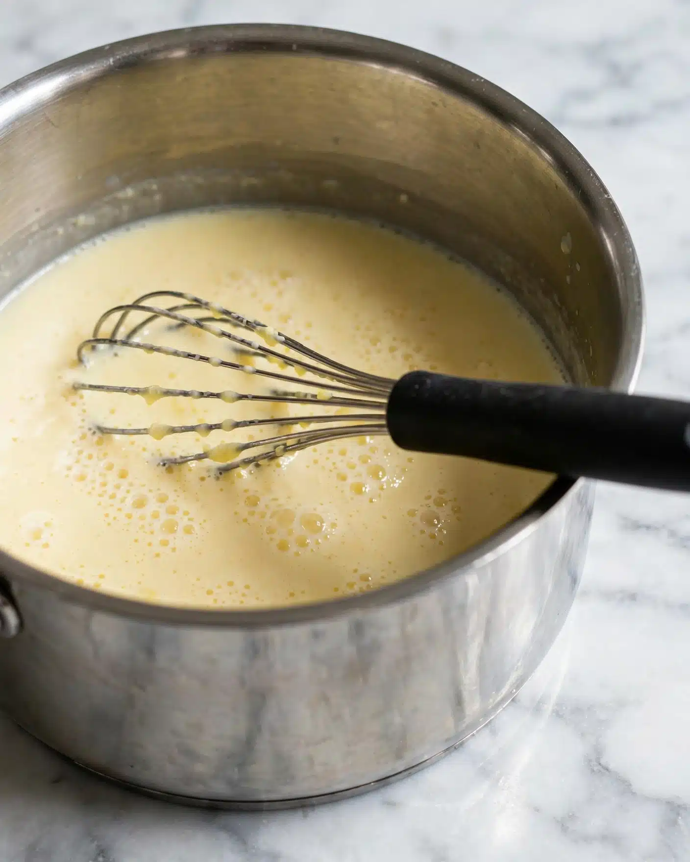 A close-up view of a shiny metal cooking pot filled with a light yellow, creamy liquid that has small bubbles on the surface. A metal whisk with a black handle rests inside the pot, partially submerged in the liquid, with the handle angled toward the right side of the frame. The pot sits on a white marbled surface that softly reflects some light. Photo taken with an iphone --ar 4:5 --v 7