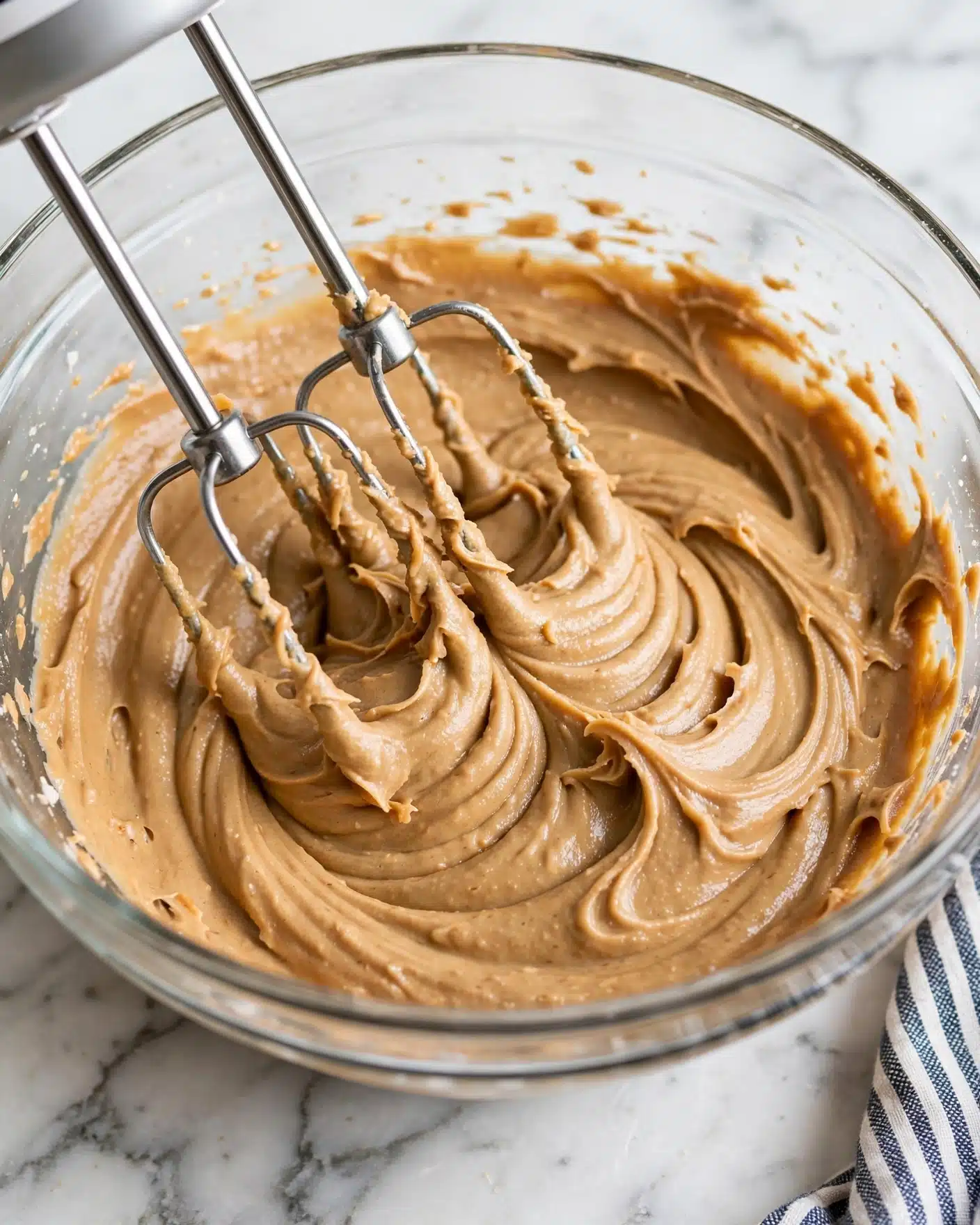 Monster Cookie Dough Dip Recipe 4 A clear glass bowl holds a thick, creamy light brown mixture with a soft, smooth texture, showing swirls and peaks from mixing. Two silver beaters from a mixer are partially embedded in the mixture, with some of it clinging to the beaters. The bowl is set on a white marbled surface with a hint of a striped cloth in the lower right corner. Photo taken with an iphone --ar 4:5 --v 7