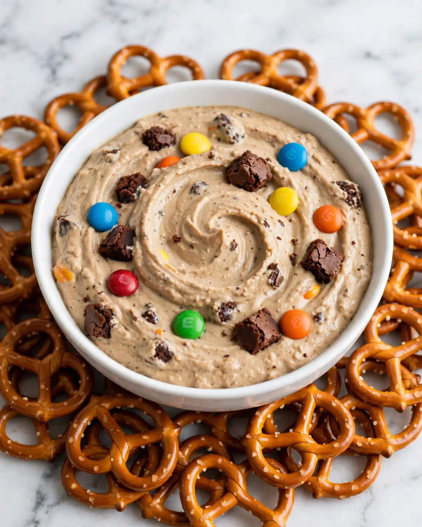 Monster Cookie Dough Dip Recipe 5 A white bowl is full of light brown dip that looks creamy and thick, mixed with colorful round candy pieces in blue, red, yellow, green, and orange, along with small dark brown chunks. The bowl sits on a pile of shiny, golden brown pretzels that form a ring around it. The background is a white marbled surface. photo taken with an iphone --ar 4:5 --v 7