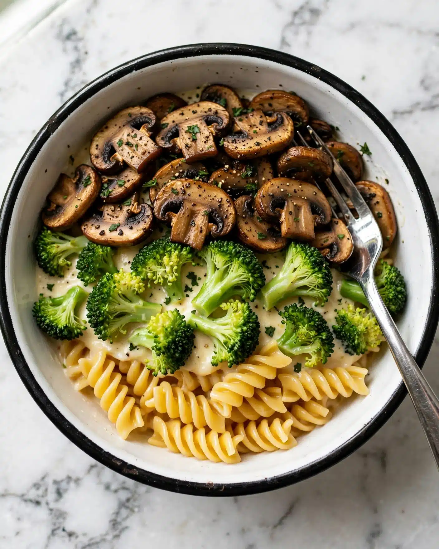 A white bowl with a black rim holds a creamy pasta dish with three main layers visible: the bottom layer is ribbed spiral pasta with a golden-yellow color, followed by bright green broccoli florets scattered evenly on top, and the top layer consists of browned mushroom slices with some black pepper and small green herb bits sprinkled all over. A shiny metal fork is partially inside the bowl, resting among the pasta and vegetables, all shown on a white marbled surface. photo taken with an iphone --ar 4:5 --v 7