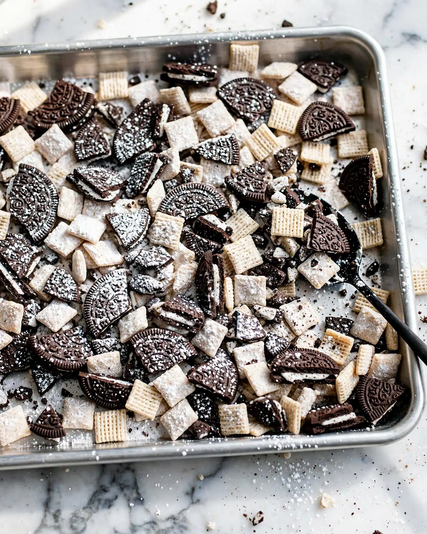 A close-up of a baking tray filled with a mix of black and dark brown broken cookie pieces, mostly sandwich cookies with white cream, and small white square cereal pieces. Both the cookies and cereal are covered with a light dusting of fine white powdered sugar, giving the entire mix a slightly frosted look. The tray is silver and rests on a white marbled surface. A thin black spoon is partially visible on the right side, resting atop the mixture. photo taken with an iphone --ar 4:5 --v 7