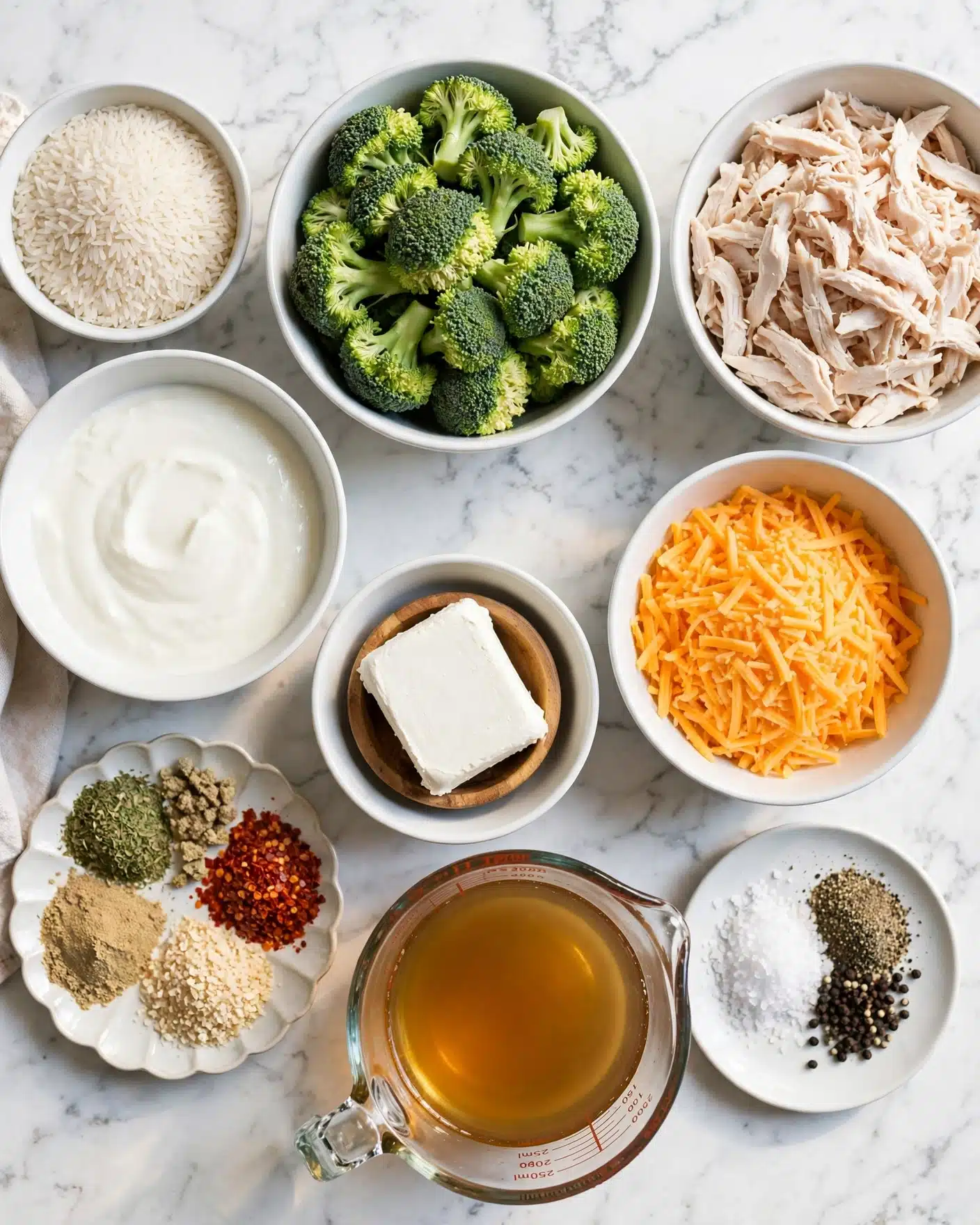 A top-down view of several white bowls and a glass measuring cup arranged on a white marbled surface, containing raw white rice in a small bowl, bright green broccoli florets filling a large bowl, shredded pale cooked chicken in another large bowl, a bowl of shredded orange cheddar cheese, a bowl of smooth white yogurt, a small scalloped plate with four green, red, and beige dry spices arranged in sections, a glass measuring cup filled with amber-colored broth, a small bowl holding a solid block of white cream cheese with a small wooden bowl beneath it, and a small plate with coarse salt and black pepper side by side, all displayed neatly and clearly; photo taken with an iphone --ar 4:5 --v 7