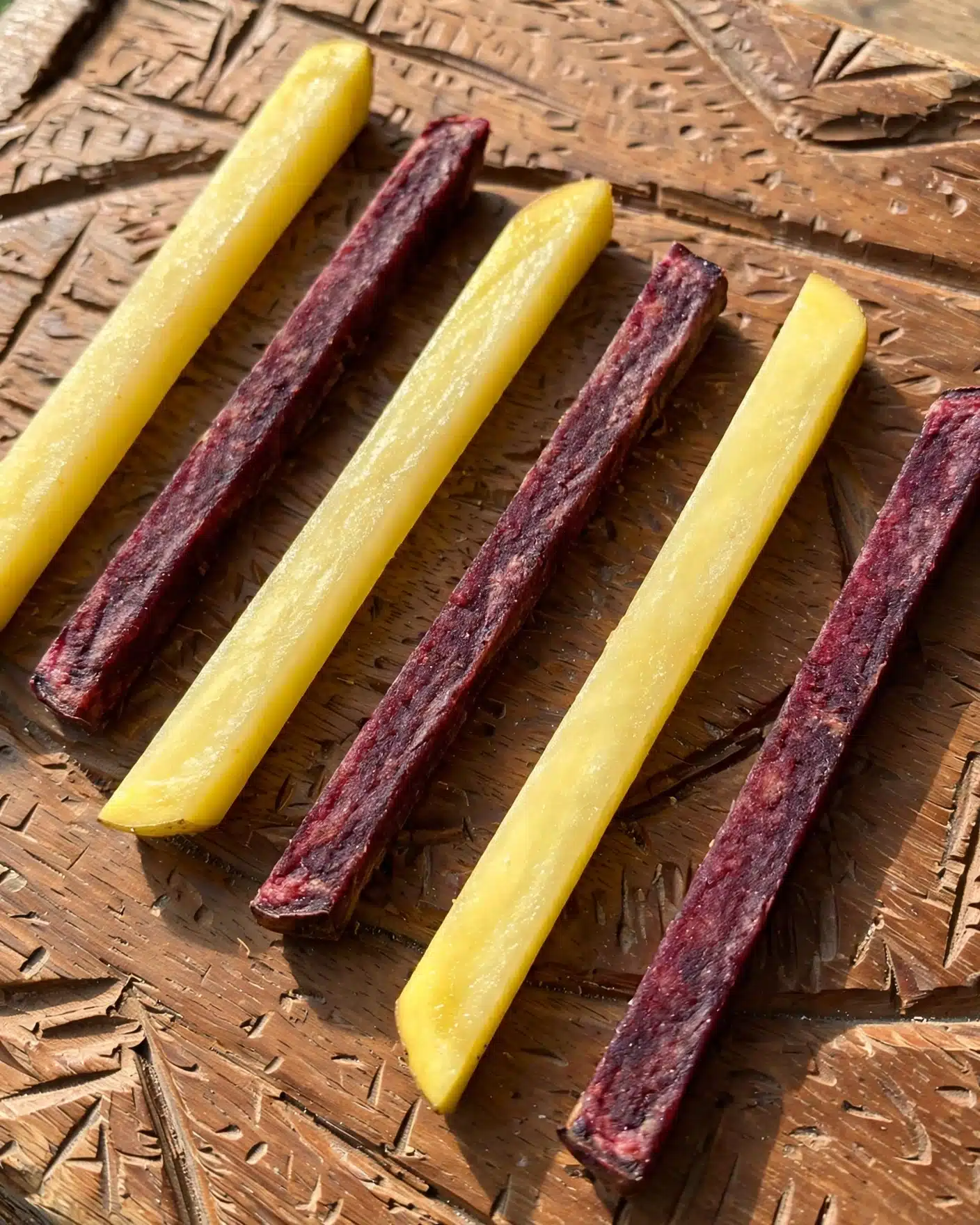The image shows six potato sticks laying flat on a brown wooden surface with a carved texture. The sticks alternate colors, with three pale yellow sticks showing smooth, slightly glossy surfaces and three dark reddish-purple sticks that have a more matte texture and rough edges. Each stick is roughly the same size and shape, long and rectangular with angled ends. The light in the image casts soft shadows to the lower right, adding depth and detail to the textures. Photo taken with an iphone --ar 4:5 --v 7