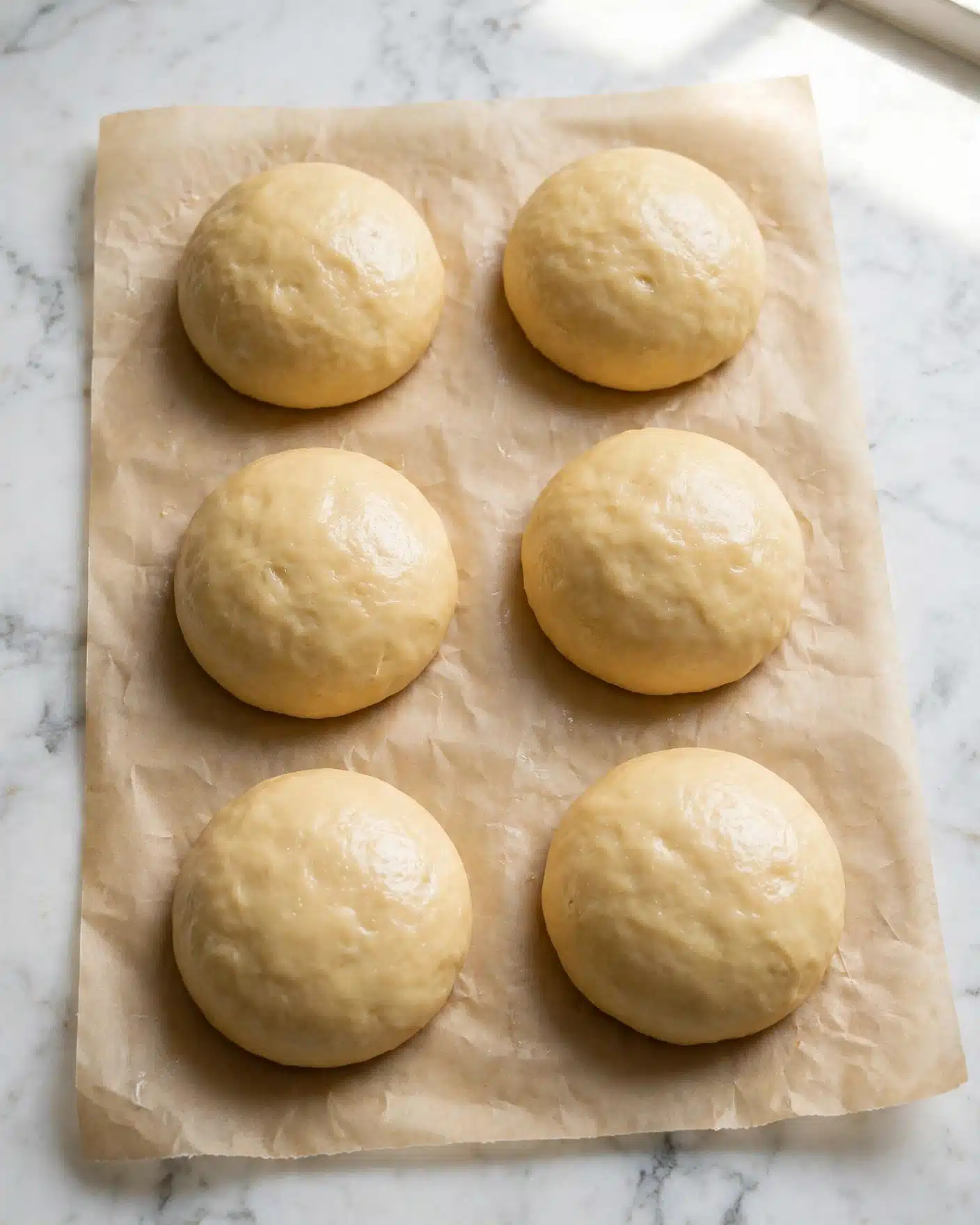 The image shows six round dough balls evenly placed on a sheet of parchment paper. Each dough ball has a smooth, pale golden surface with a slightly shiny texture, spaced apart in two vertical columns of three on a white marbled background. The dough balls look soft and slightly raised, ready for baking. photo taken with an iphone --ar 4:5 --v 7