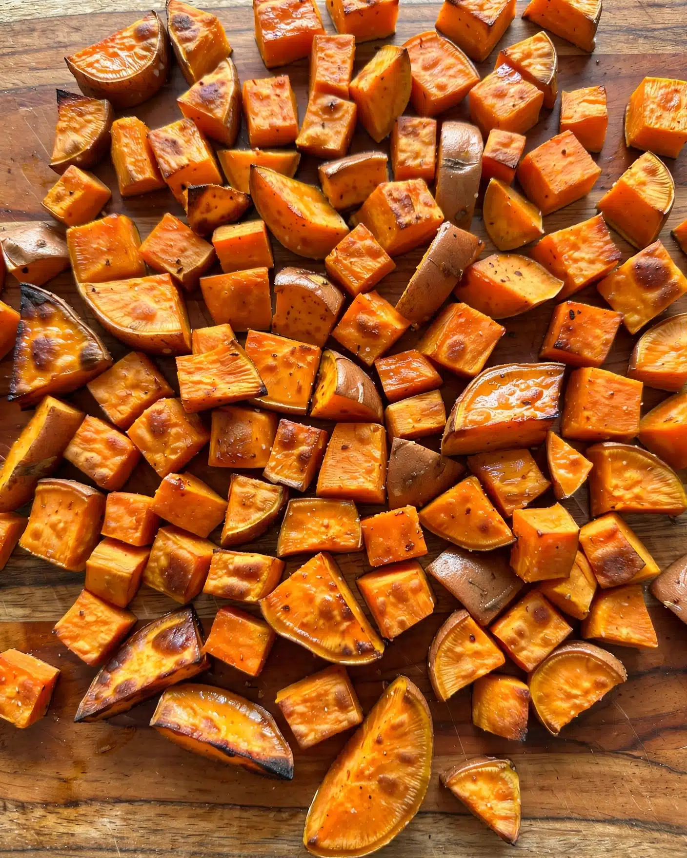 Sweet Potato Burger Bowl Recipe 4 The image shows many orange sweet potato pieces on a wooden board. The sweet potatoes are cut into small, thick cubes and some wedge shapes. The pieces have a smooth, slightly shiny texture and are spread out close to each other. The wooden board has a warm brown color with visible grain lines. There is no other object in the image. Photo taken with an iphone --ar 4:5 --v 7