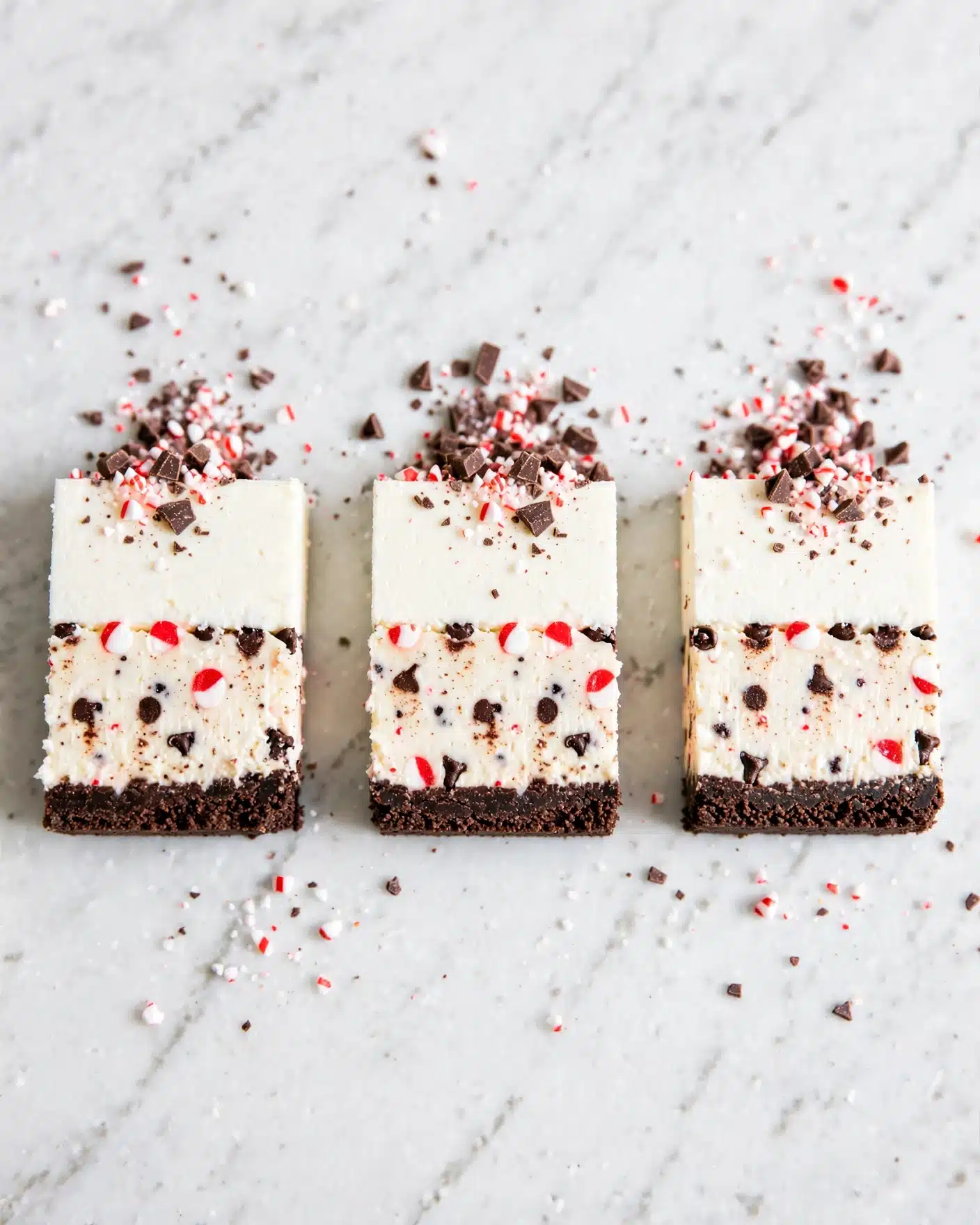 Peppermint Bark White Chocolate Mousse Cheesecake Recipe 5 The image shows three slices of a dessert arranged in a row on a white marbled surface. Each slice has three clear layers: a dark, crumbly base at the bottom, a thick middle layer that is creamy white with small red and dark pieces mixed in, and a smooth top layer of white cream. The top cream layer is decorated with small bits of crushed peppermint and chocolate shavings. Photo taken with an iphone --ar 4:5 --v 7