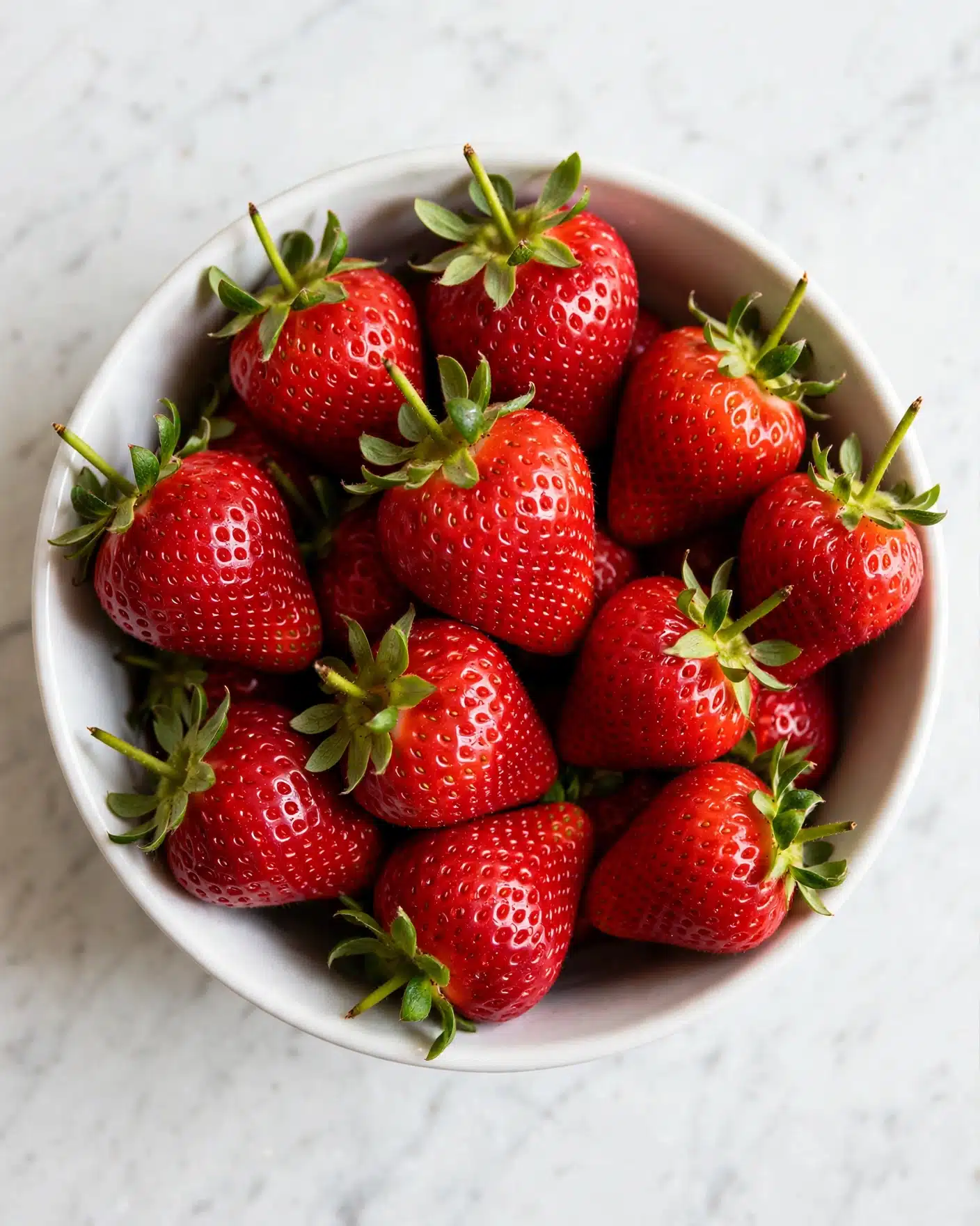 A close-up image of a white bowl filled with bright red strawberries with green leaves still attached. The strawberries look fresh and shiny with detailed texture on their surface. The bowl is placed on a white marbled surface, adding a clean and simple background that highlights the vibrant colors of the strawberries. photo taken with an iphone --ar 4:5 --v 7