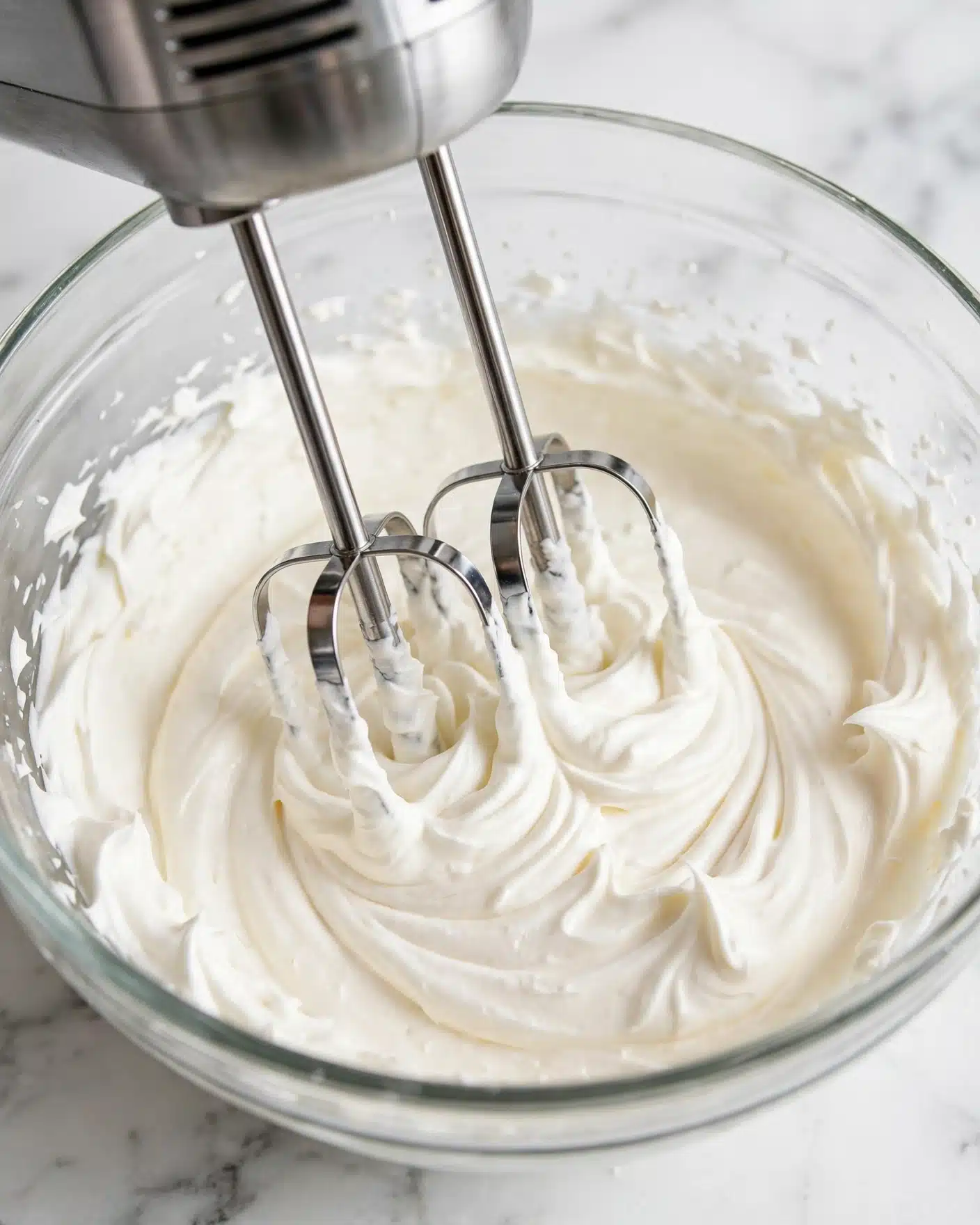 Keto Cream Cheese Mints Recipe 4 In this close-up image, a metal mixer with two silver beaters is seen blending a thick, creamy, white mixture in a clear glass bowl. The creamy mixture has a smooth texture with soft peaks formed around the edges of the bowl and clinging to the beaters. The background shows a white marbled texture, adding a clean and bright contrast to the soft cream. photo taken with an iphone --ar 4:5 --v 7