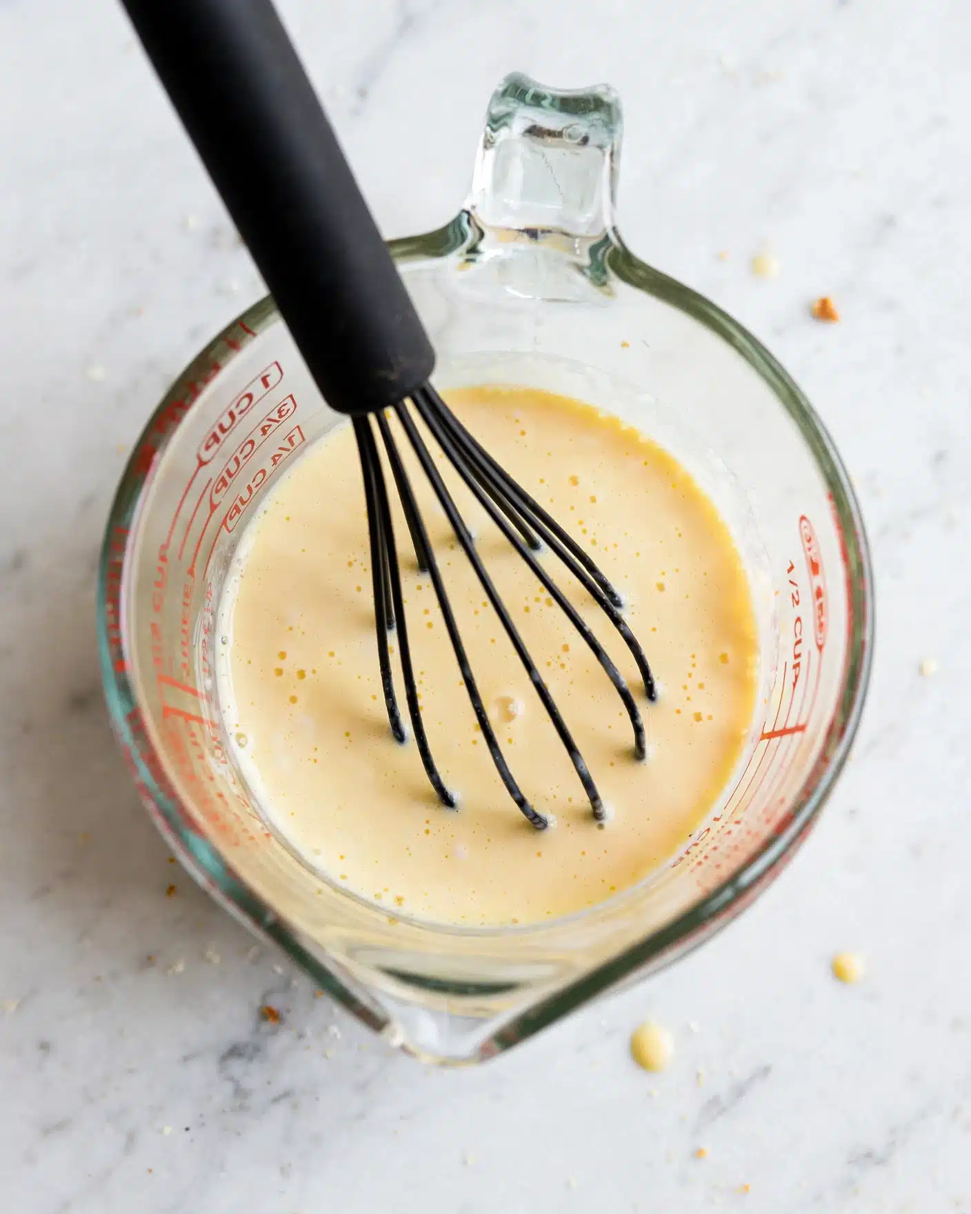 A clear glass measuring cup holds a light yellow liquid mixture. Inside, a black whisk with a smooth handle stirs the mixture, which appears creamy and smooth with small bubbles on the surface. The cup sits on a white marbled surface, and part of the measuring cup’s red measurement marks are visible on the side. Photo taken with an iphone --ar 4:5 --v 7