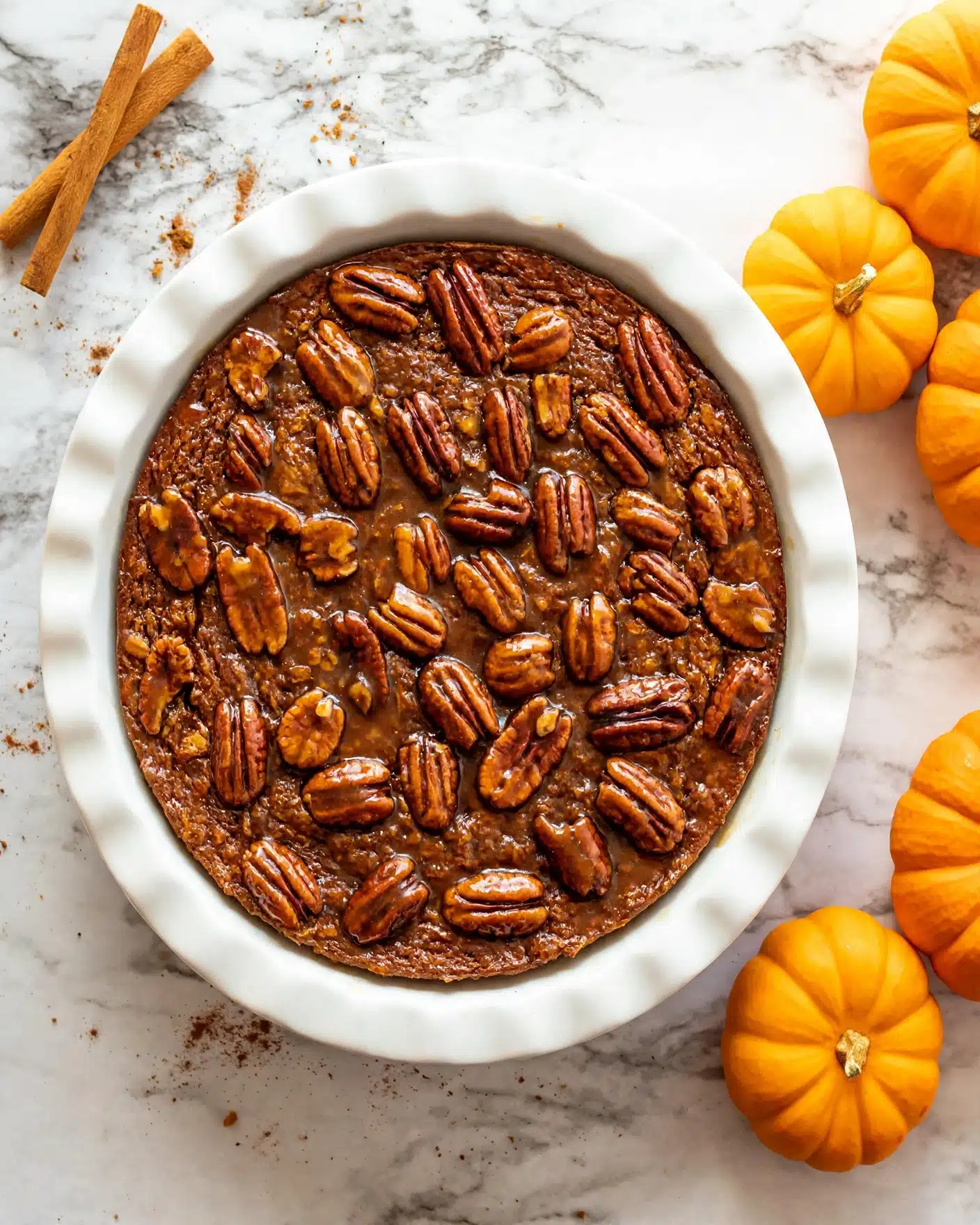 Pumpkin Spice Baked Oatmeal Recipe 5 The image shows a round pecan pie in a white ceramic dish. The pie has one thick layer with a rich brown color and a slightly rough texture. On top of the pie, there is an even spread of whole pecan halves that are glossy and a darker brown, arranged close to each other, covering the surface. The pie dish sits on a white marbled surface, and to the right of the dish are several small bright orange pumpkins that add a warm, autumn feel. Photo taken with an iphone --ar 4:5 --v 7
