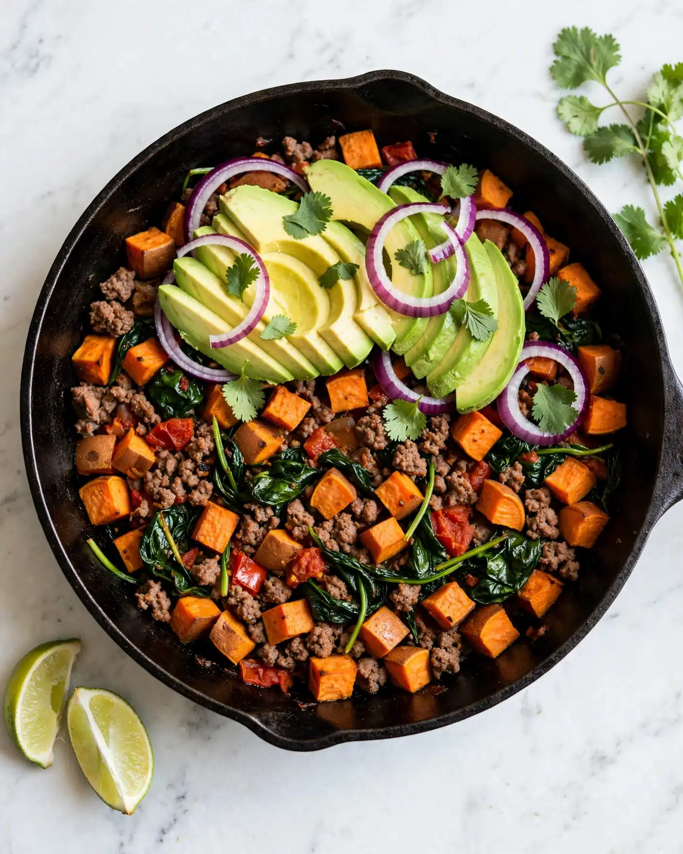 The image shows a close-up of a cooked dish in a black pan containing small cubes of orange sweet potatoes, browned ground meat, wilted dark green spinach leaves, and bits of red tomato. On top are several slices of bright green avocado fanned out, with thin rings of purple-red onion and a few fresh green cilantro leaves scattered around. The pan rests on a white marbled surface with lime wedges and cilantro sprigs partially visible nearby, creating a fresh and colorful presentation. photo taken with an iphone --ar 4:5 --v 7