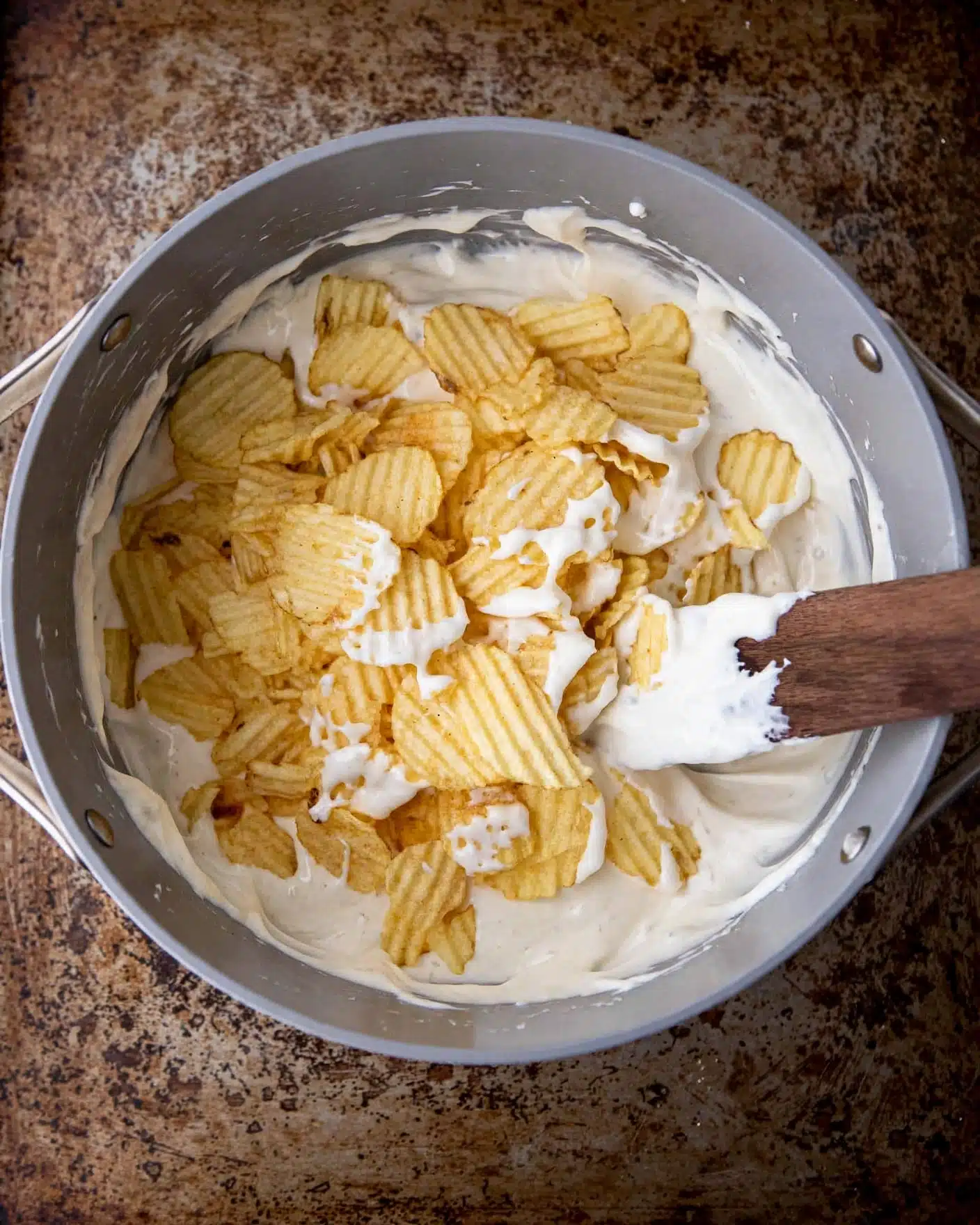 Ruffles Chips Marshmallow Treats Recipe 4 A gray pot sits on a rustic brown surface filled with a mix of two layers: the bottom layer is a thick, creamy white sauce that partially coats the sides of the pot, and the top layer is made of yellow ridged potato chips scattered loosely. A wooden spatula rests inside, partially covered with the white sauce, positioned on the right side of the pot. photo taken with an iphone --ar 4:5 --v 7