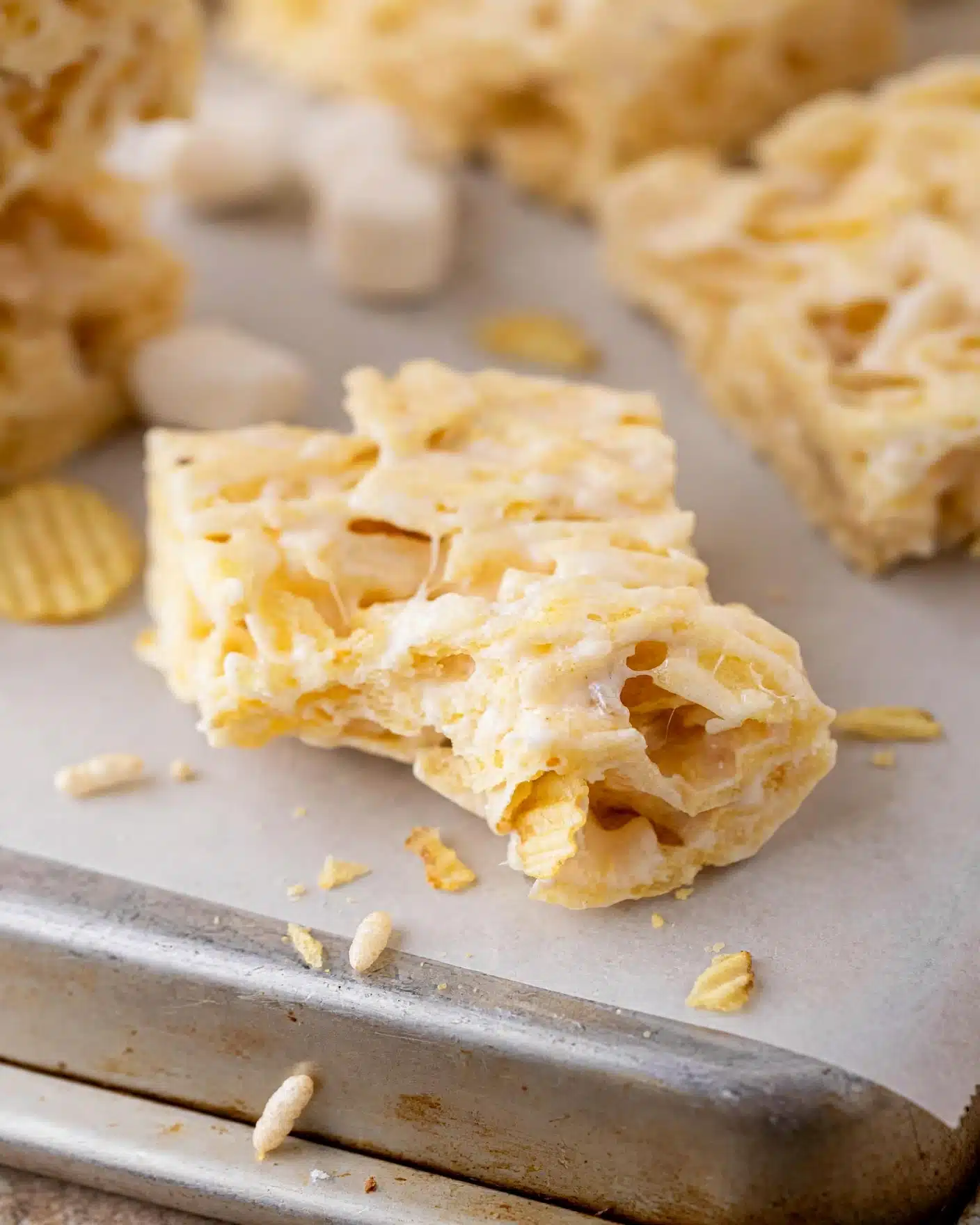 Ruffles Chips Marshmallow Treats Recipe 5 The image shows a close-up of a light golden square crispy rice treat with a smooth, slightly shiny texture, resting on white parchment paper on a metal tray. The treat has visible layers of puffed rice cereal fused together with a creamy, sticky marshmallow layer, giving a chewy and airy look. There are more similar squares blurred in the soft white marbled textured background. Photo taken with an iphone --ar 4:5 --v 7