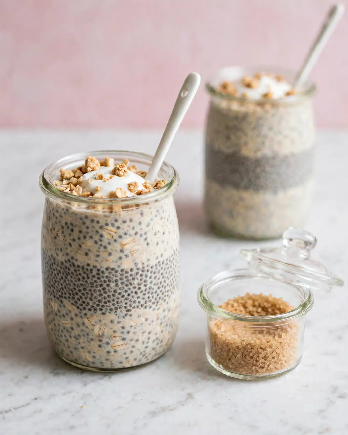 Coffee Overnight Oats Recipe 5 The image shows a clear glass jar filled with three thick layers of a creamy chia seed and oat mix, speckled with dark chia seeds throughout. The top layer is finished with a small dollop of white yogurt or cream and sprinkled with light brown crumbled nuts or granola, creating a crumbly texture. The jar is placed on a white marbled surface with a small clear glass container of brown sugar next to it. In the background, there is a second jar with the same contents slightly blurred. A thin white spoon sticks out from the first jar, set against a soft pink backdrop. Photo taken with an iphone --ar 4:5 --v 7
