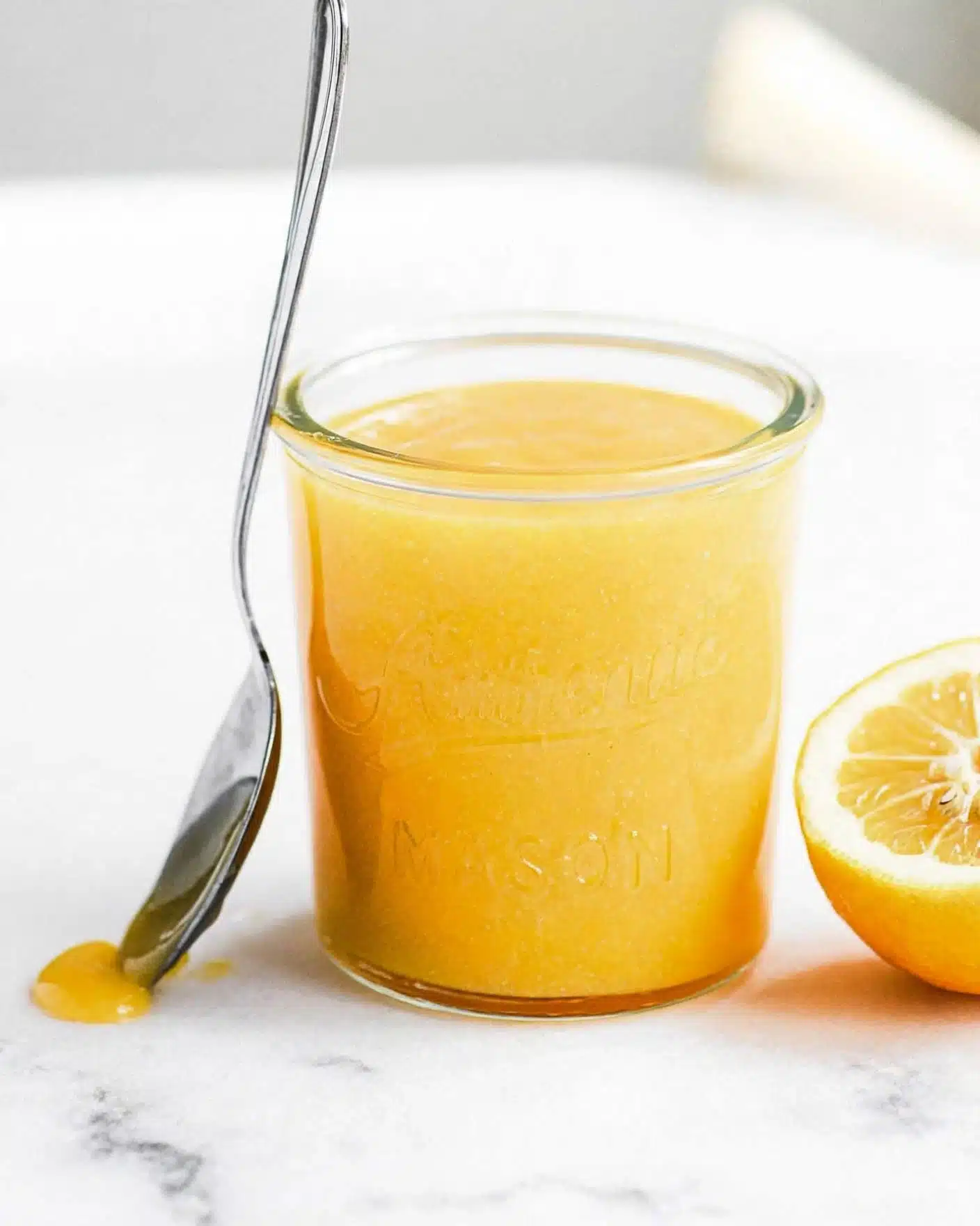 Lemon Poppy Seed Bars Recipe 4 A clear glass jar filled with smooth, bright yellow lemon curd, showing a thick, creamy texture. The jar has the word
