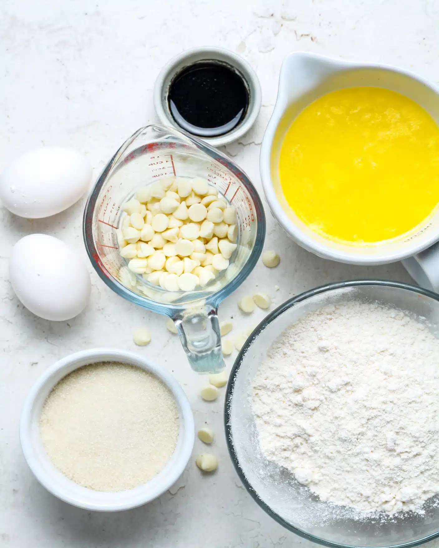 The image shows six main items arranged on a white marbled surface: two white eggs on the left side, a small white bowl filled with light beige sugar below the eggs, a clear glass measuring cup filled with white chocolate chips in the center, a small white bowl with dark black vanilla extract above the chocolate chips, a white bowl with melted yellow butter on the right side, and a large clear glass bowl with white flour below the butter bowl. All items are spaced out and photographed from above. Photo taken with an iphone --ar 4:5 --v 7