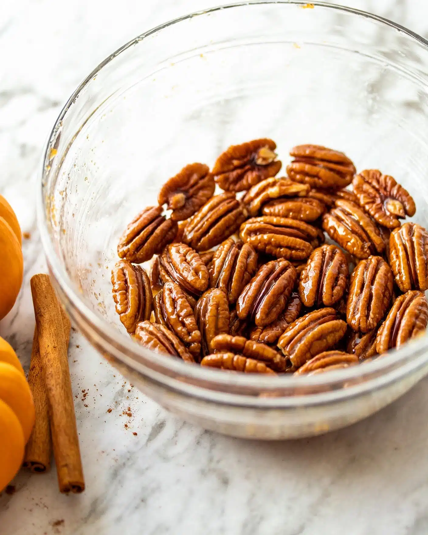 Pumpkin Spice Baked Oatmeal Recipe 4 A clear glass bowl filled with a single layer of glazed pecans that have a shiny, textured surface showing their bumpy, curved shapes in a warm golden brown color, sitting on a white marbled surface. To the left of the bowl, a cinnamon stick rests on the surface near two small orange pumpkins, adding a warm, cozy feel to the scene. The pecans are layered closely together, covering the bottom of the bowl fully, and the glass bowl allows light to pass through, highlighting the pecans' rich color and glossiness. photo taken with an iphone --ar 4:5 --v 7