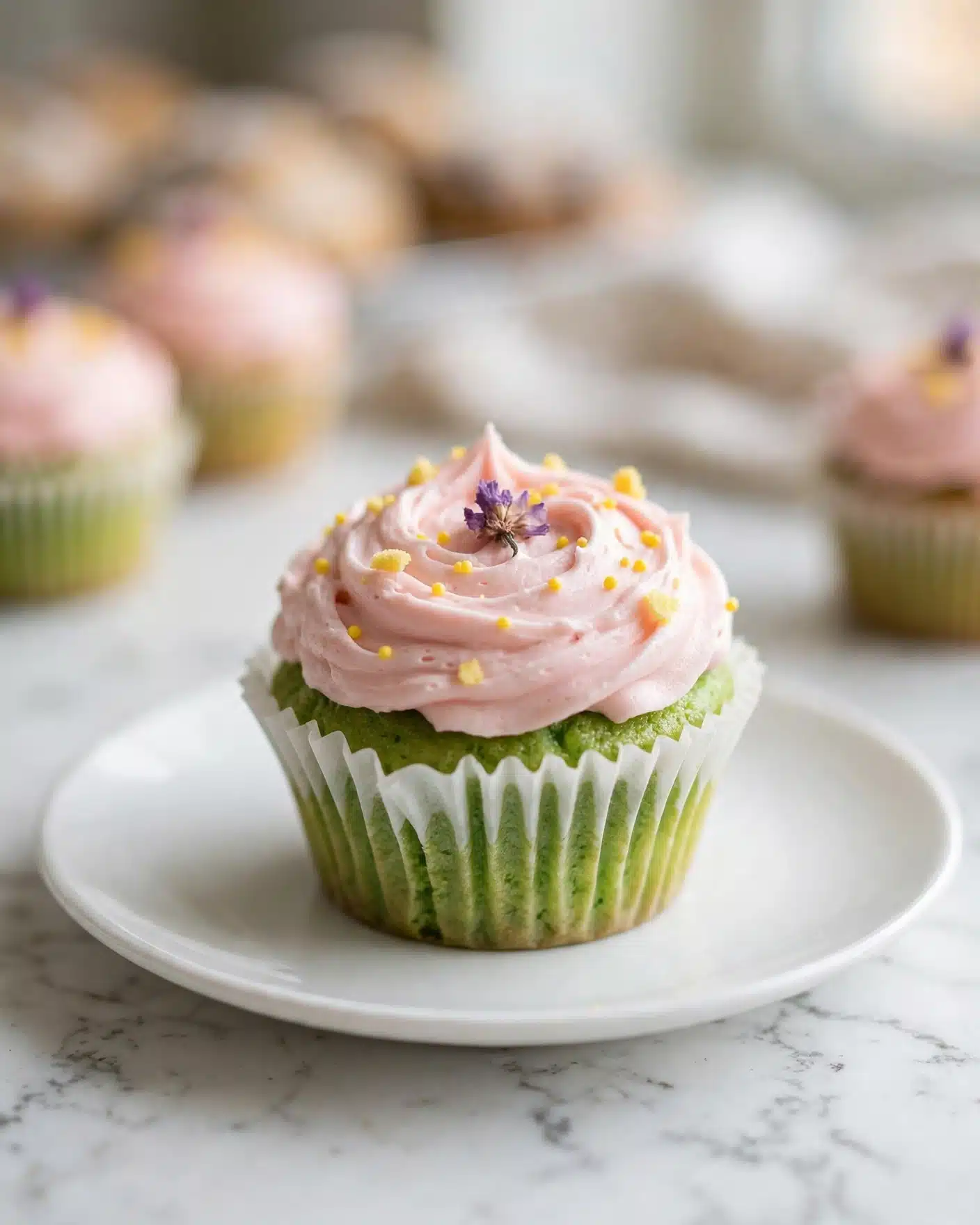 The image shows a single cupcake set on a white marbled surface, placed on a white plate. The cupcake has two layers: the bottom layer is a bright green, moist cake sitting inside a white pleated paper liner, and the top layer is a swirl of smooth, light pink frosting with a soft texture. The frosting is decorated with tiny yellow bits scattered on top and a small purple dried flower placed in the middle. The background is softly blurred, hinting at more similar cupcakes. Photo taken with an iphone --ar 4:5 --v 7