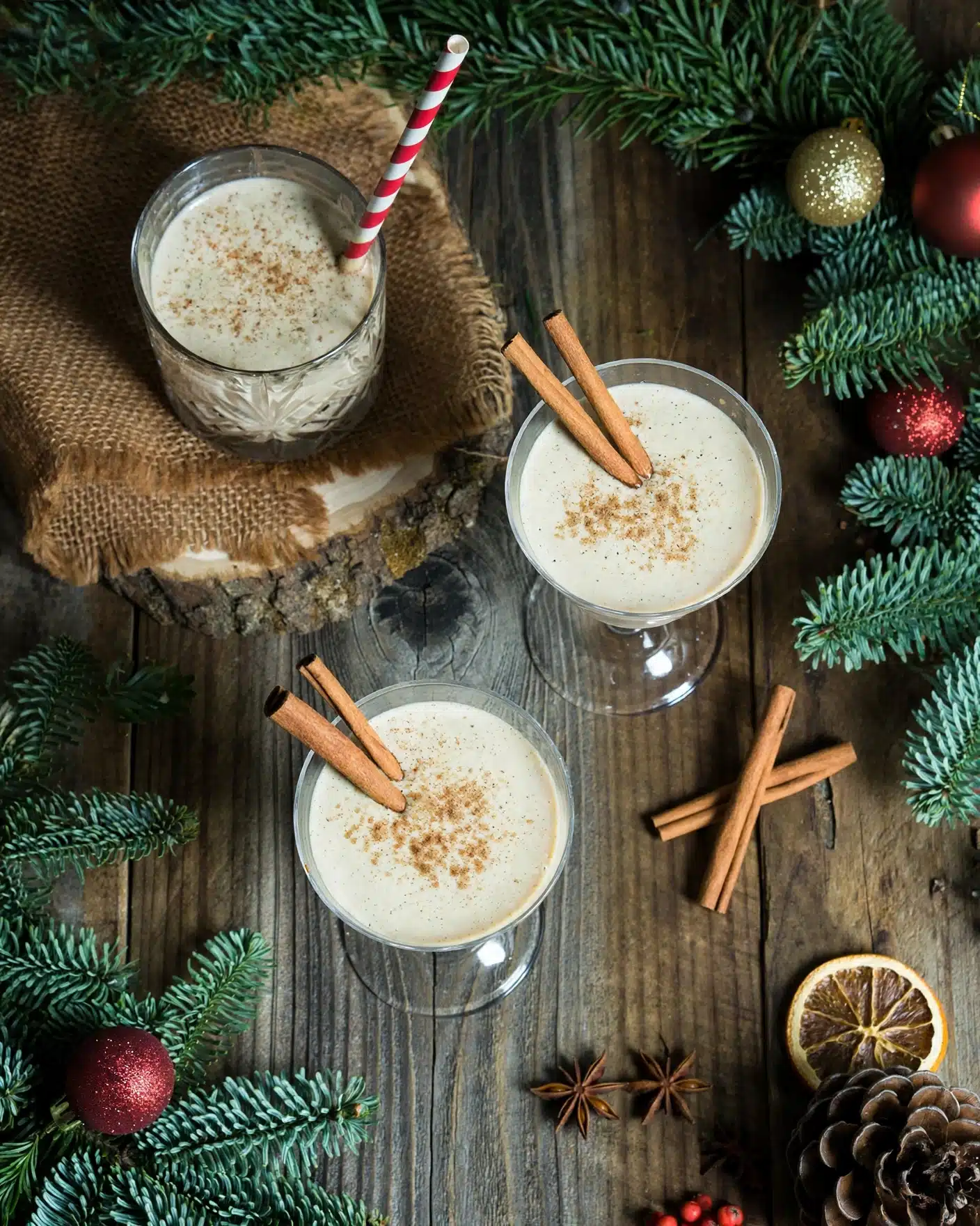 Jamie Oliver's Eggnog Recipe 5 Three glasses of creamy light beige eggnog are placed on a dark wooden table with visible grain and knots. Two glasses have a cinnamon stick on the rim, and all have a sprinkle of brown nutmeg or cinnamon powder on top. One glass has a red and white striped paper straw. One glass is set on a rough brown burlap cloth over a tree bark slice, creating a rustic feel. Around the glasses, there are green pine branches, red berries, shiny red and gold Christmas ornaments, and a large pine cone, giving a festive holiday atmosphere. The photo taken with an iphone --ar 4:5 --v 7