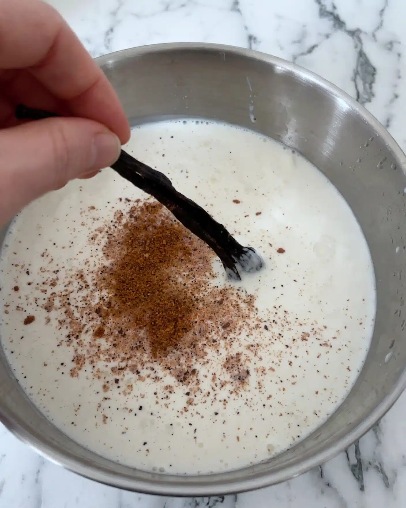 Jamie Oliver's Eggnog Recipe 4 The image shows a close-up of a white bowl filled with a thick, creamy white liquid that looks like milk or cream. On the liquid's surface, there is a light brown powder sprinkled in the middle, likely cinnamon or a similar spice. Above the liquid, a dark brown or black vanilla bean pod is held horizontally by a woman's hand, slightly blurred, with its tip dipping into the liquid. The bowl rests on a white marbled surface, and the photo is focused on the vanilla pod and the sprinkled powder. Photo taken with an iphone --ar 4:5 --v 7