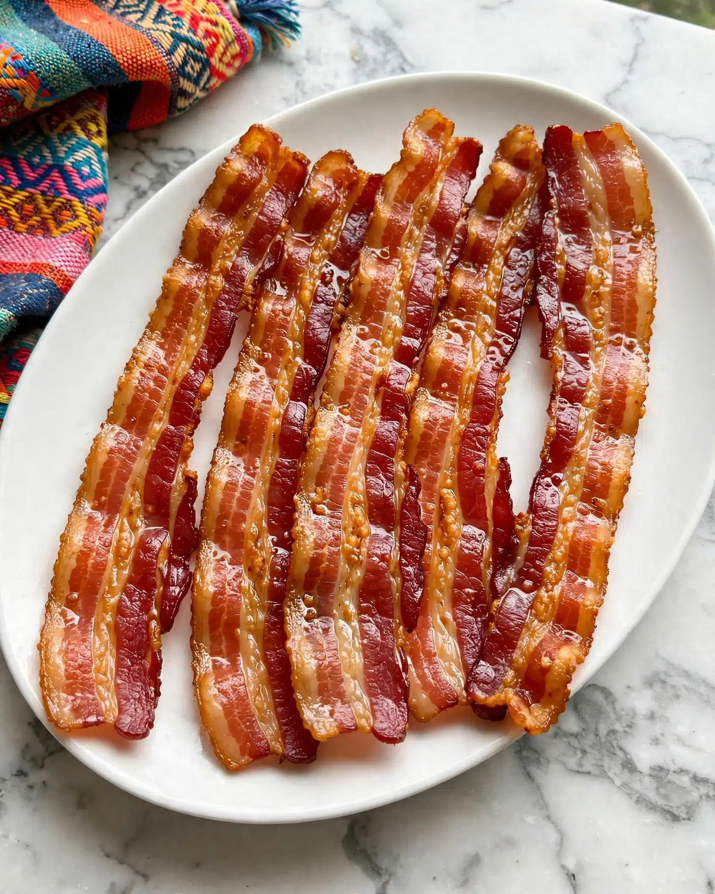 Brown Sugar Bacon Recipe 5 A white oval plate holds five long, shiny strips of cooked bacon. The bacon strips have a bright reddish-brown color with some lighter, crispy golden edges. The glossy surface of each strip shows a slight oily texture that makes them look freshly cooked. The plate sits on a white marbled surface with a colorful patterned cloth at the upper left corner, adding a touch of brightness. The bacon strips are arranged side by side, overlapping a bit in the center, clearly showing their different shades and textures. Photo taken with an iphone --ar 4:5 --v 7