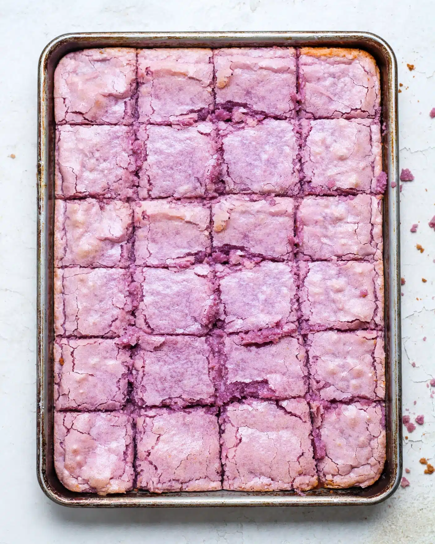 This image shows a close-up view of a tray of square-shaped pink brownies with a cracked surface. The brownies have a shiny, slightly rough top layer, with some darker pink, moist-looking edges where the pieces meet. The texture looks soft and dense inside, visible at the cracks and broken edges. The squares are arranged tightly side by side on a white marbled surface. photo taken with an iphone --ar 4:5 --v 7