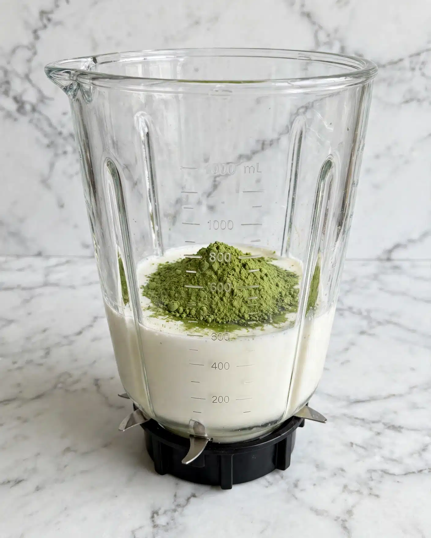 Matcha Mille Crepe Cake Recipe 4 Inside a clear blender jar with measurement marks, there is a layer of white liquid at the bottom mixed with a pile of green powder sitting on top, giving a contrast between the smooth liquid and the fine powder. The blender blades are visible around the base of the container, and the background shows a white marbled texture. photo taken with an iphone --ar 4:5 --v 7