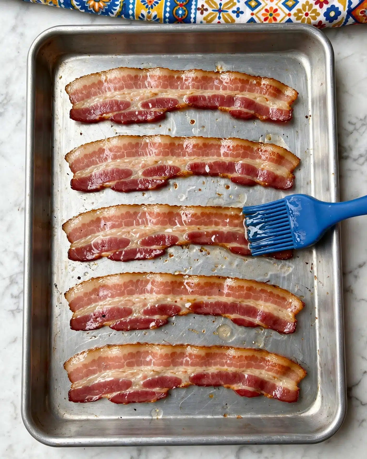 Brown Sugar Bacon Recipe 4 The image shows five strips of cooked bacon laid out in a single layer on a metal baking tray. The bacon has pinkish-red and light cream fat colors with a slightly shiny texture due to grease. A blue silicone brush is brushing the bacon on the right side of the third strip from the top. The background is a white marbled texture with part of a brightly patterned cloth visible at the top edge. Photo taken with an iphone --ar 4:5 --v 7