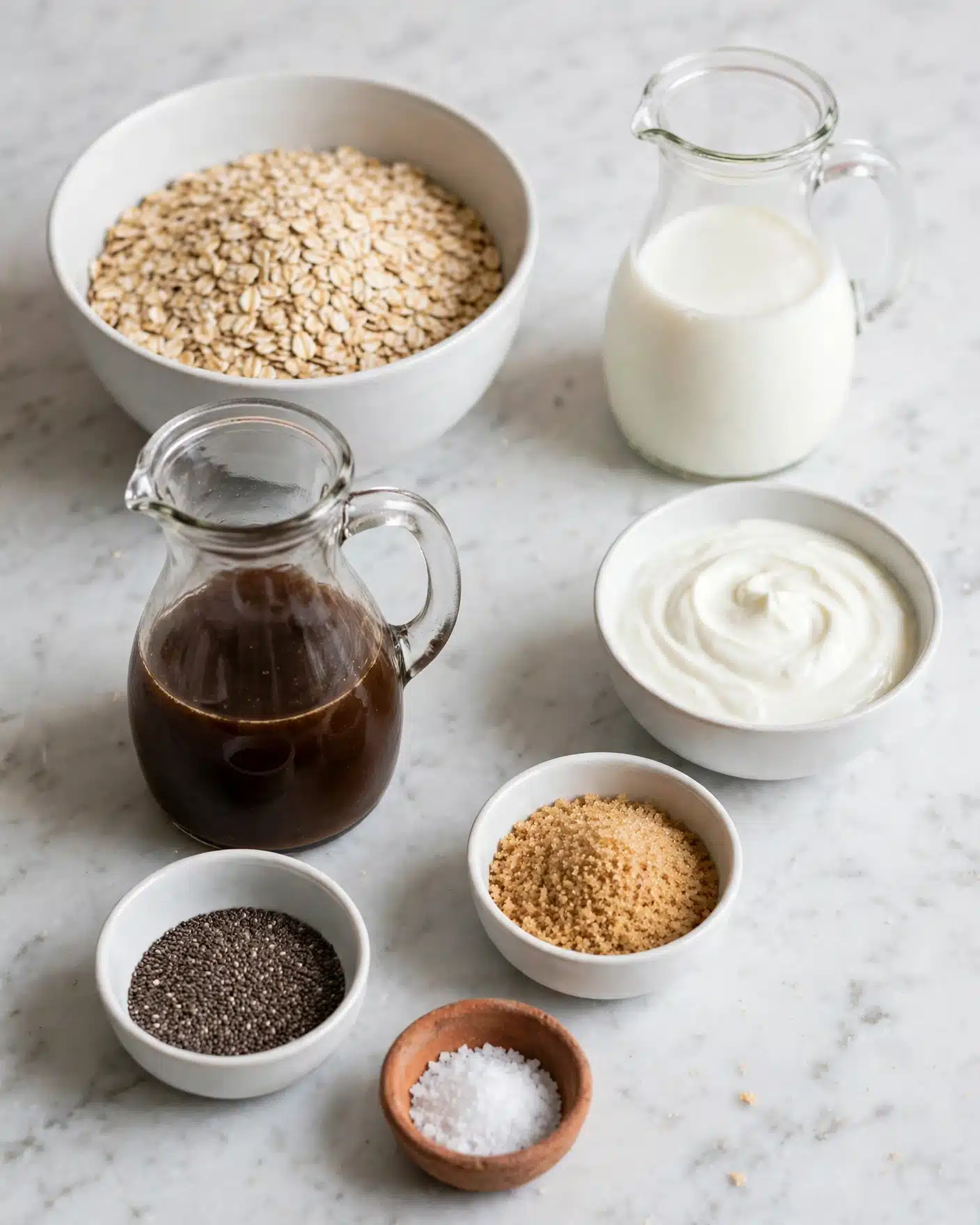 Coffee Overnight Oats Recipe 4 The image shows six small bowls and jugs arranged on a white marbled surface. The largest bowl on the left is white and filled with light brown rolled oats, with a rough texture. To the right, a clear glass jug holds white milk, smooth in texture. Below this, a smaller white bowl contains thick and creamy white yogurt, arranged in gentle swirls. To the left, a glass jug has a dark brown liquid that looks smooth and dense. Below this jug, two small white bowls hold tiny black chia seeds on the left and light brown granulated brown sugar on the right. At the bottom, there is a tiny clay bowl with a small amount of coarse white salt. All containers are neatly spaced, creating a clean and natural setup. photo taken with an iphone --ar 4:5 --v 7
