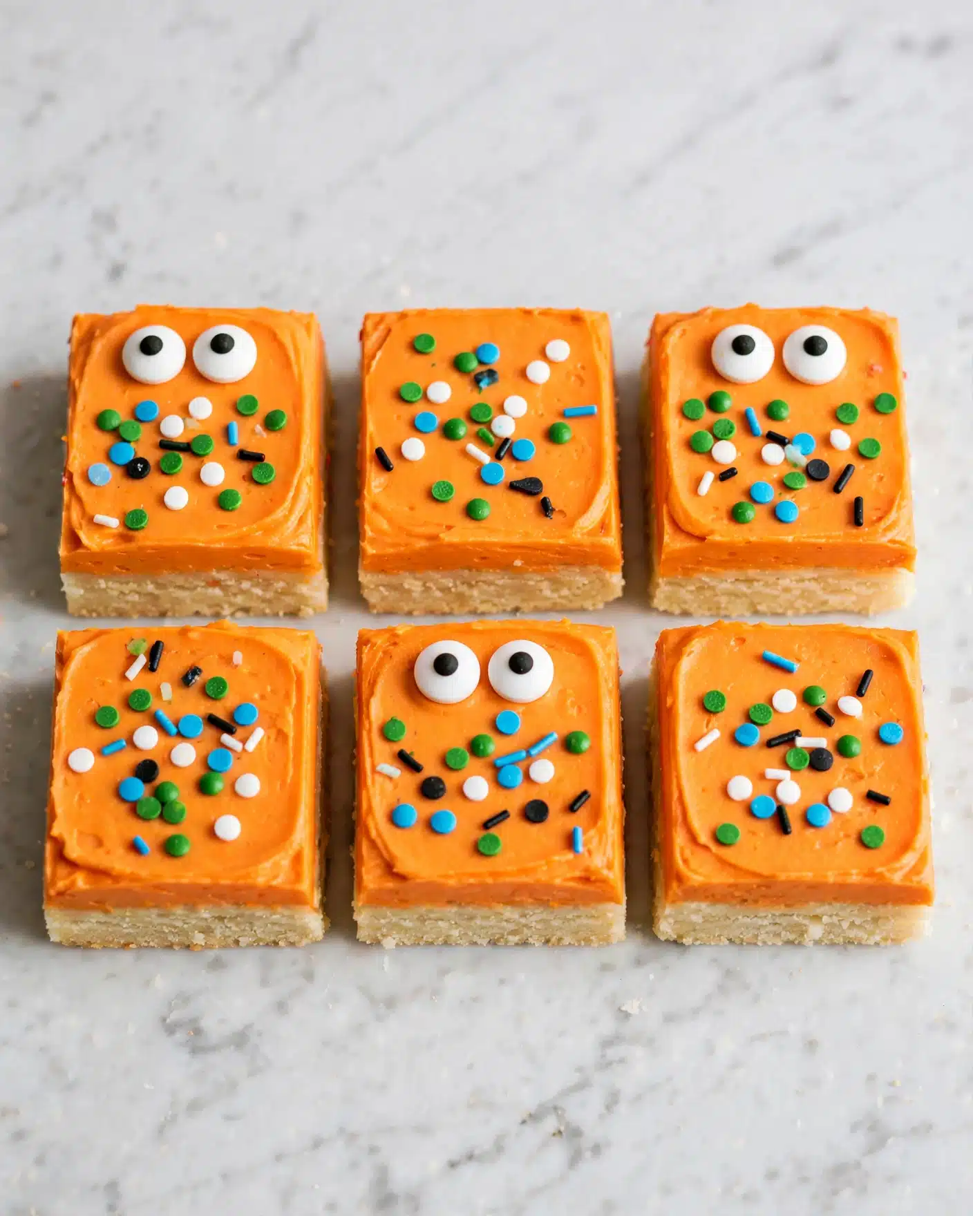 Halloween Sugar Cookie Bars Recipe 5 A close-up view of square treats arranged side by side, each with two layers: a light-colored bottom layer and a thick, smooth bright orange top layer. On the orange layer, there are colorful small sprinkles in green, blue, white, and black scattered around. Some squares have two white candy eyes with black centers placed near the edges, giving a playful look. The treats are set on a wooden surface but should be imagined on a white marbled background. photo taken with an iphone --ar 4:5 --v 7