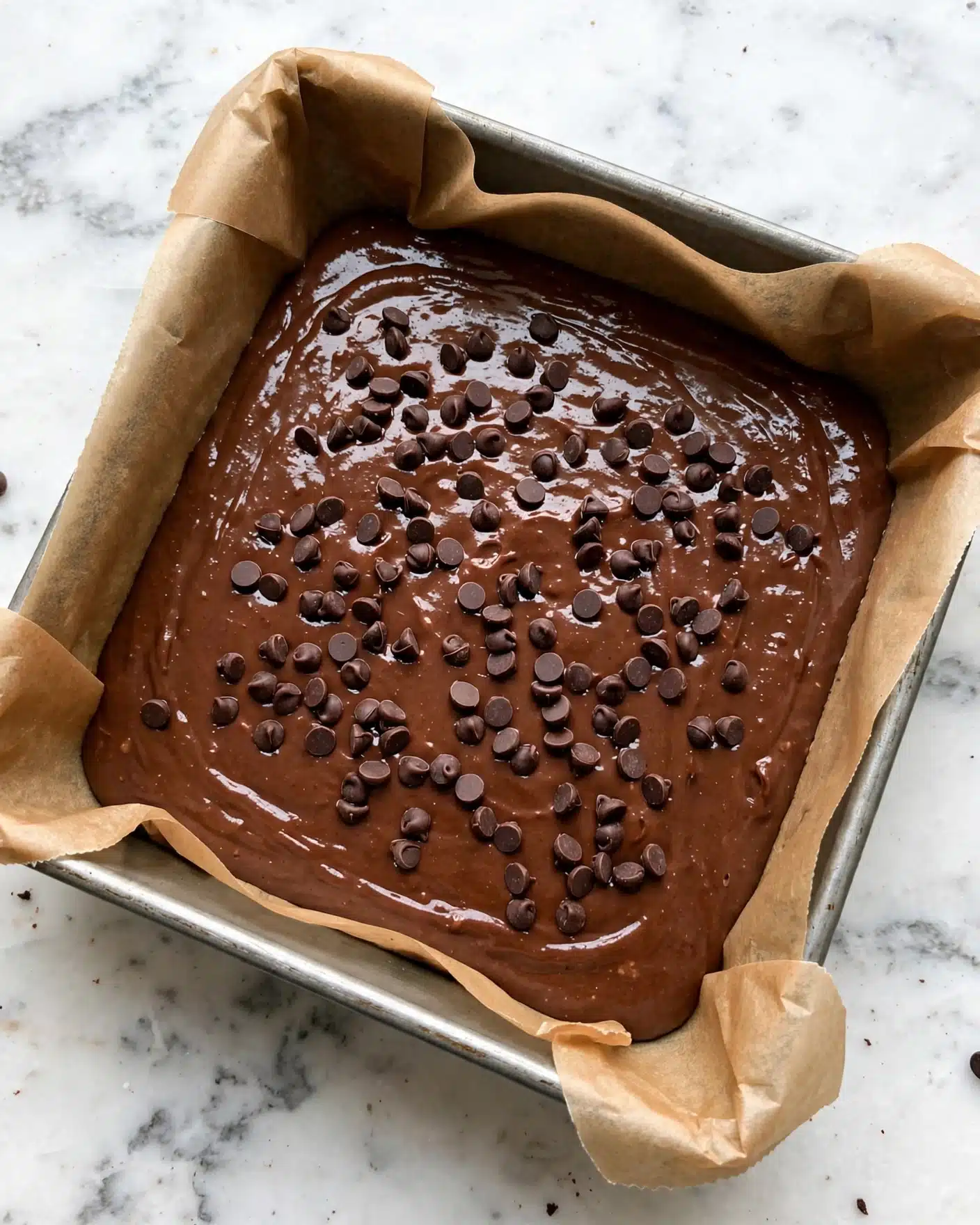 A square baking pan lined with brown parchment paper holds a single layer of smooth, rich chocolate batter spread evenly across the bottom. The top layer is decorated with scattered small and medium dark chocolate chips that create a textured look against the glossy chocolate surface. The pan sits on a white marbled background, showing clear detail of the pan’s edges and the parchment paper folding up the sides. photo taken with an iphone --ar 4:5 --v 7