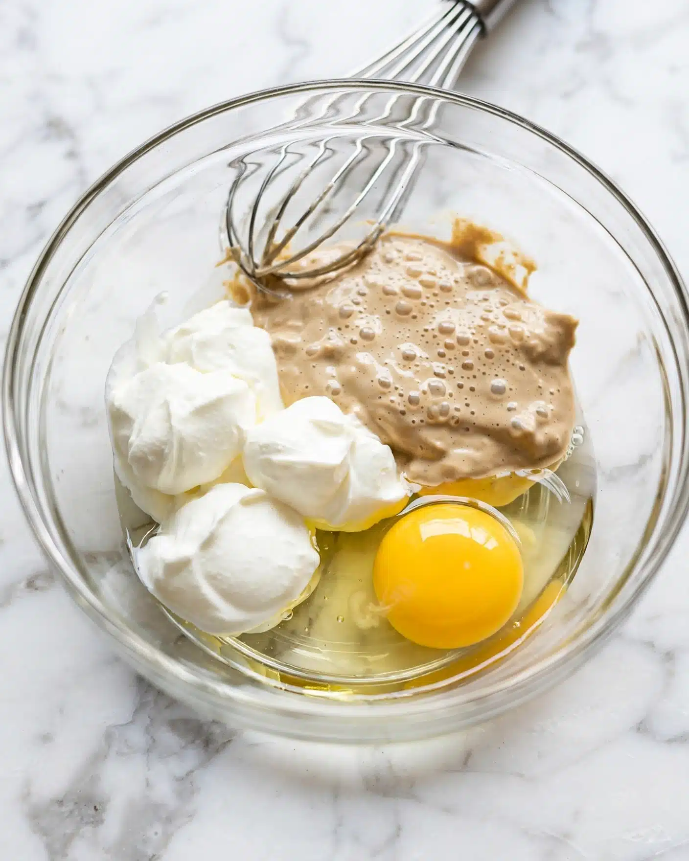A clear glass bowl is placed on a white marbled surface, containing a mix of ingredients that lay in separate layers but are about to be mixed. At the bottom is a clear runny liquid, likely oil or water, next to a bright yellow whole egg yolk towards the front right. Around the yolk, there are dollops of thick white cream or yogurt on the left side and a light tan creamy substance with small bubbles on top toward the upper right. A metal whisk is positioned inside the bowl, resting on the ingredients near the back. The overall scene is bright and clean, showing the beginning of a recipe mix. Photo taken with an iphone --ar 4:5 --v 7