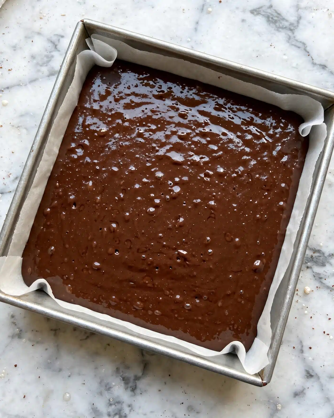 Cookie Dough Brownies: Fudgy Brownies Topped with Edible Cookie Dough and Colorful M&M’s Recipe 4 A square metal baking pan lined with white parchment paper is filled with a smooth, shiny layer of dark brown chocolate batter. The batter spreads evenly across the pan with small bubbles and tiny texture variations visible on its surface. The pan is placed on a white marbled texture surface. photo taken with an iphone --ar 4:5 --v 7