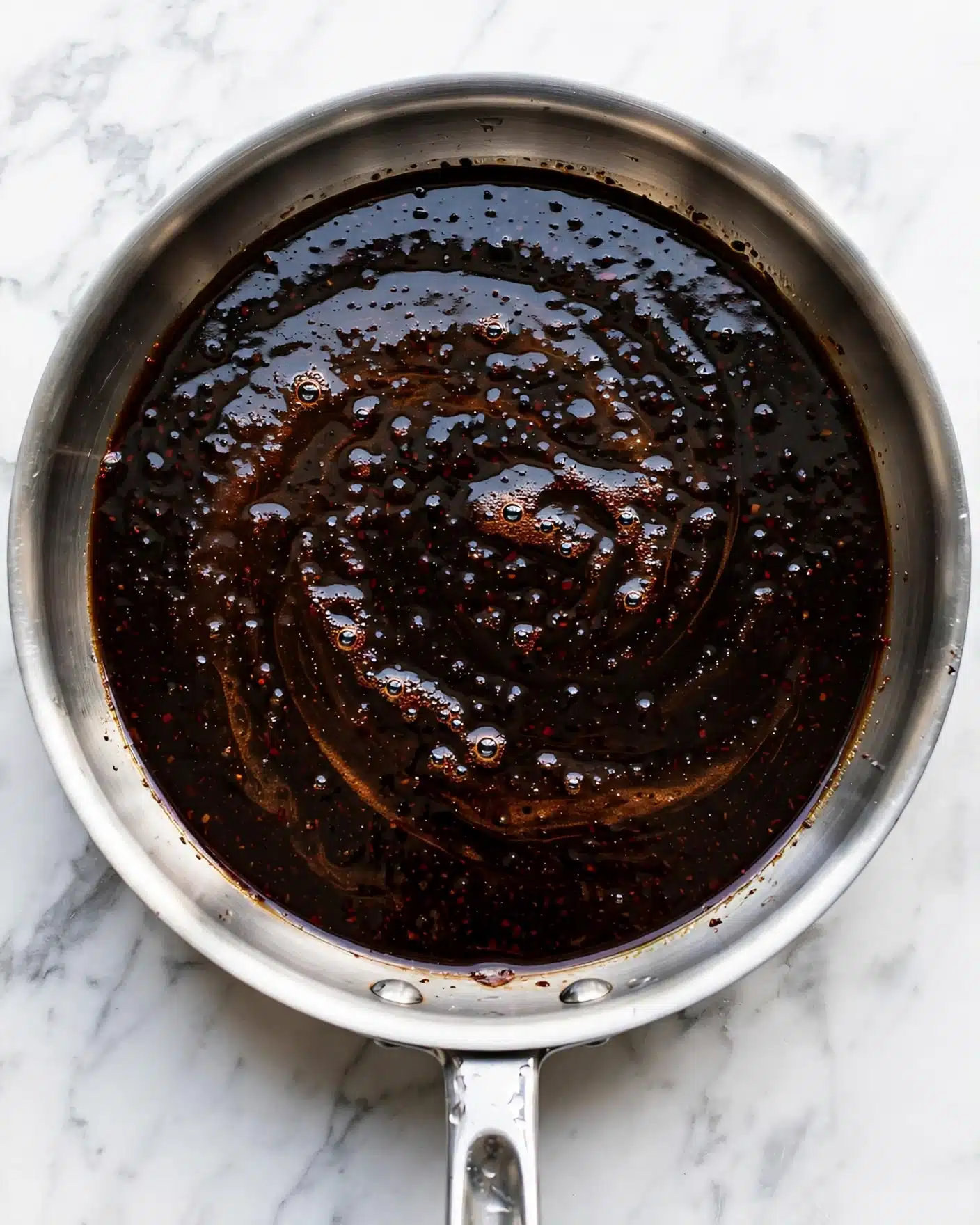 A close-up top view of a single layer of a dark liquid with a shiny, slightly bubbly and smooth surface inside a shiny silver pan, placed on a white marbled surface. The liquid is almost black with hints of brown and red tones reflecting light, creating a subtle swirl pattern. The pan handle is visible at the bottom center of the image. photo taken with an iphone --ar 4:5 --v 7