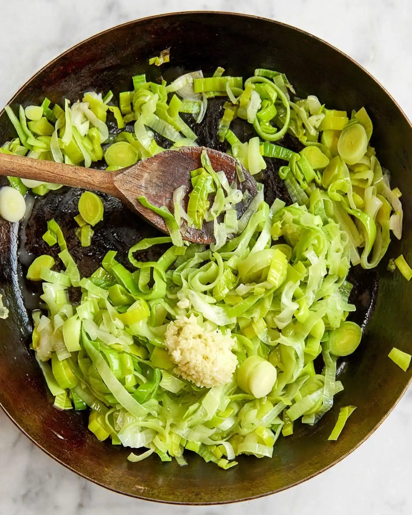 A close-up view of a dark pan filled with many thin slices of green leeks cooked or cooking, with a small pile of light yellow minced garlic on top near the center. A wooden spoon is resting inside the pan, holding some leeks, positioned in the upper left area. The background is a white marbled surface. photo taken with an iphone --ar 4:5 --v 7