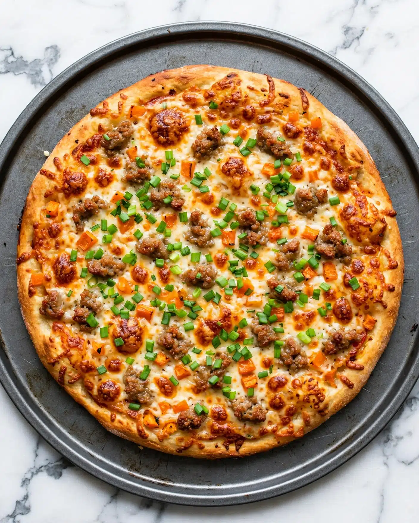 A pizza with a golden brown crust placed on a round gray pizza pan sitting on a white marbled surface. The pizza has one visible layer of melted cheese topped with small pieces of browned meat, orange bits of cooked vegetables, and a generous sprinkle of chopped green onions scattered evenly across the top, giving a fresh green contrast to the warm cooked colors below. The cheese beneath has a slight browning in some places showing a baked texture. Photo taken with an iphone --ar 4:5 --v 7