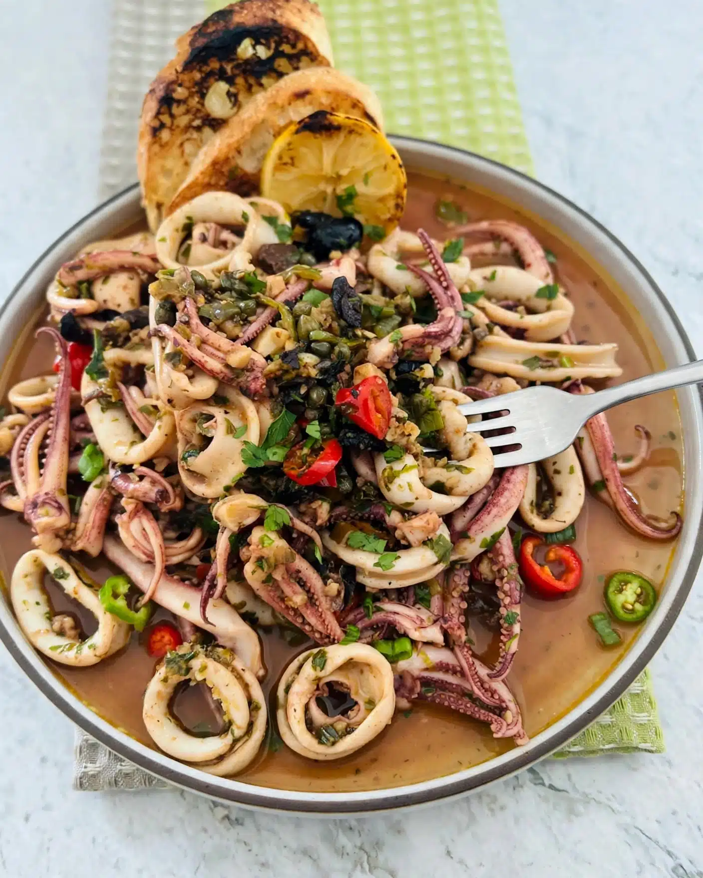 Quick and Easy Spicy Sautéed Calamari with Capers, Olives, and Herbed Breadcrumbs Recipe 5 This dish shows a white bowl full of squid rings and tentacles in a light brown sauce. On top of the squid are chopped green herbs, capers, black olives, and small pieces of red chili peppers. Behind the squid, two toasted pieces of bread and a grilled lemon half lean against the edge of the bowl. A fork is sticking into the squid on the right side. The bowl sits on a white marbled surface with a green cloth in the background. Photo taken with an iphone --ar 4:5 --v 7
