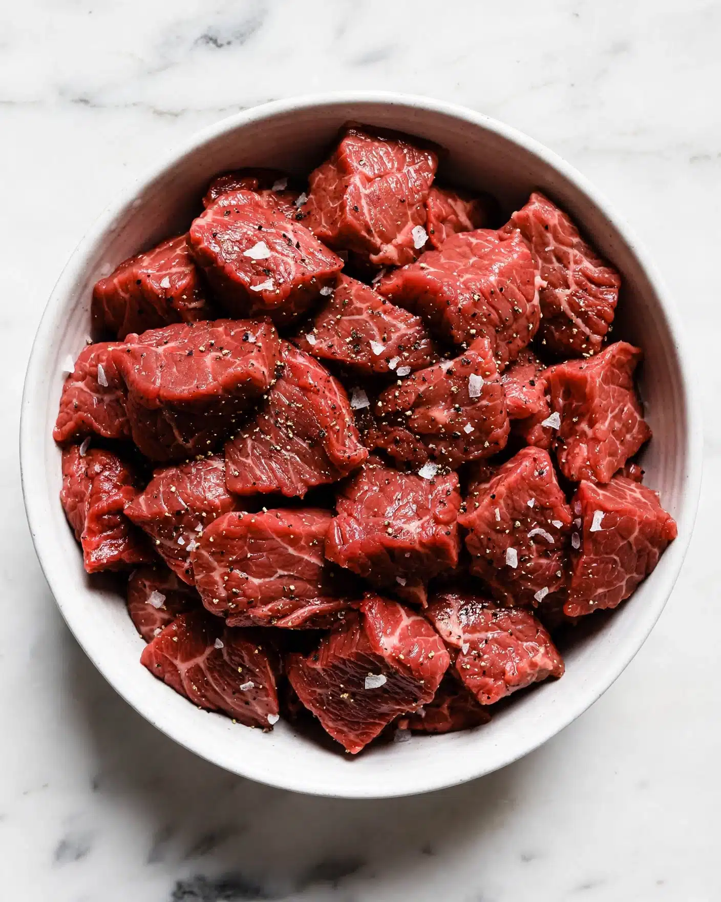 A white bowl filled with many pieces of raw red meat, each piece sprinkled with coarse salt and black pepper. The meat looks fresh and slightly shiny, with irregular shapes and varied sizes, covering the entire bowl. The bowl is placed on a white marbled surface that contrasts with the deep red of the meat, giving a clean and fresh look. photo taken with an iphone --ar 4:5 --v 7