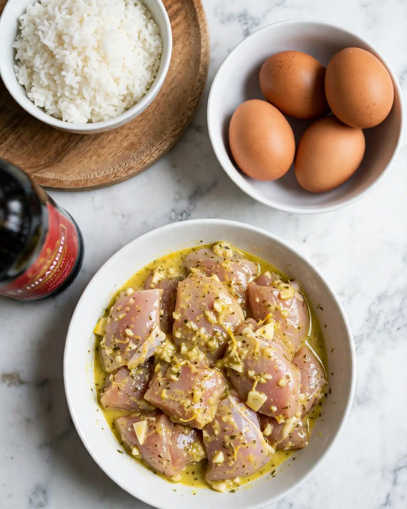 A top view of four main items arranged on a white marbled surface: a white bowl filled with raw chicken pieces marinated in a light yellow sauce sitting at the bottom center, a white bowl with four brown eggs at the top right, a white bowl with white rice resting on a round wooden board at the top left, and part of a tall dark bottle with a red label on the left side. The colors show natural food tones with soft textures for the marinated chicken, smooth eggs, and grains of rice. photo taken with an iphone --ar 4:5 --v 7