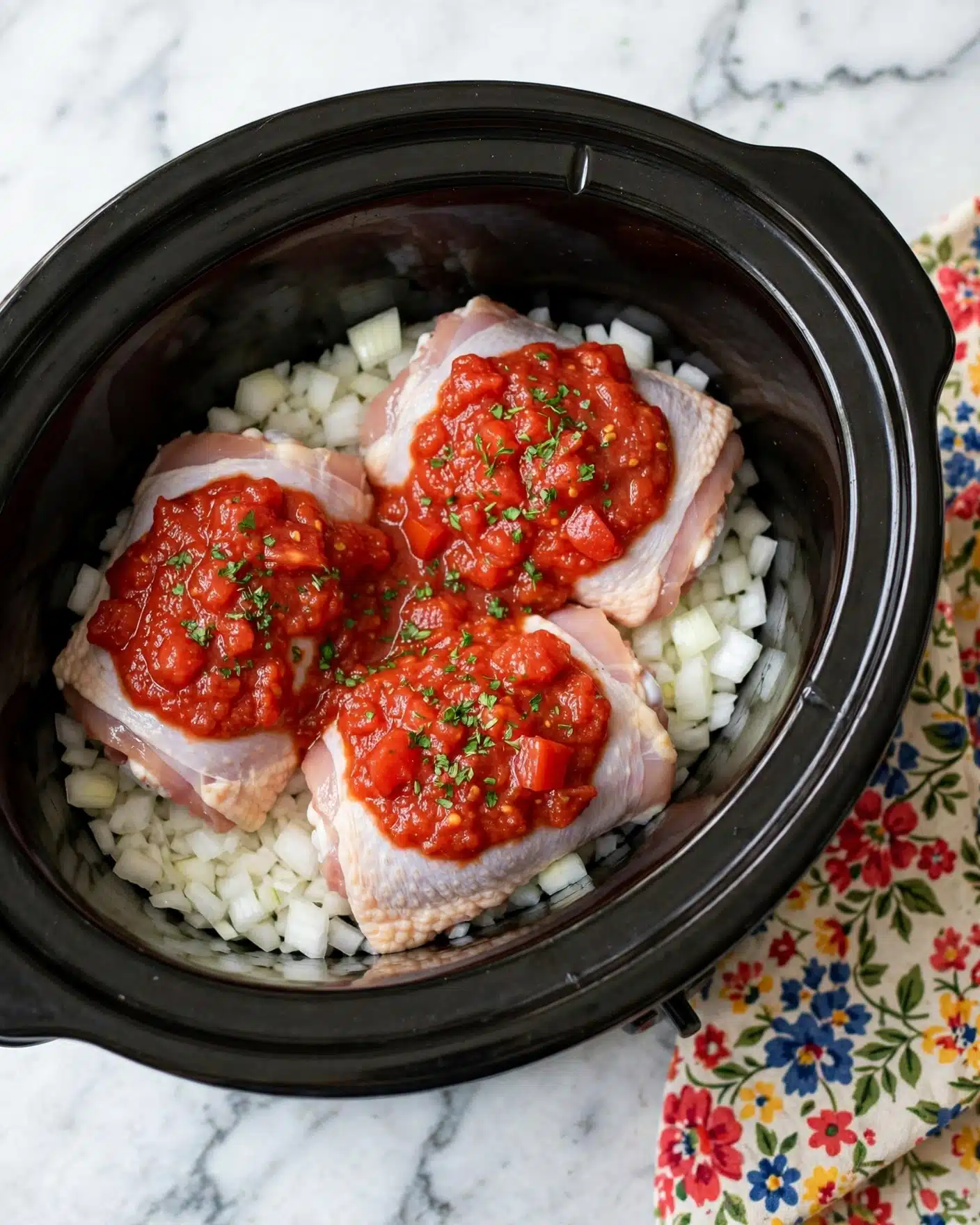 Crockpot Chicken Tacos Recipe 4 Inside a black slow cooker, there are three raw chicken thighs with light pink skin placed evenly in the center. Underneath the chicken thighs, a layer of chopped white onions covers the bottom, with small pieces spread all around. On top of the chicken thighs, there is a thick, chunky red tomato sauce with visible bits of tomato and some green herbs sprinkled on it. The entire scene sits on a white marbled surface, and a colorful cloth with red, blue, and yellow floral patterns is partially visible beside the slow cooker. Photo taken with an iphone --ar 4:5 --v 7