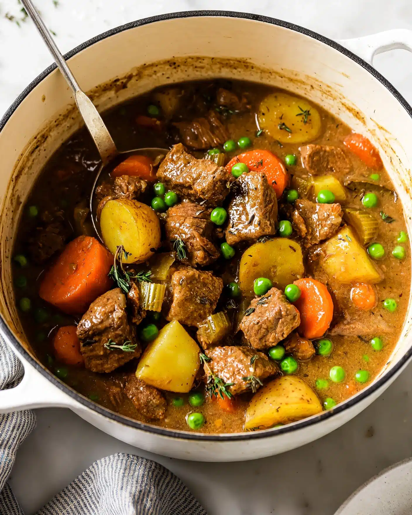 A white pot filled with thick brown stew showing chunks of dark brown beef, large pieces of bright orange carrot, yellow potatoes with peas, and pieces of celery with a pale green color, topped with small sprigs of fresh green herbs. The stew looks rich with a slightly glossy surface inside the pot, and a silver ladle is resting inside near the top left. The pot sits on a white marbled surface with a cloth napkin showing thin dark and light stripes nearby. photo taken with an iphone --ar 4:5 --v 7