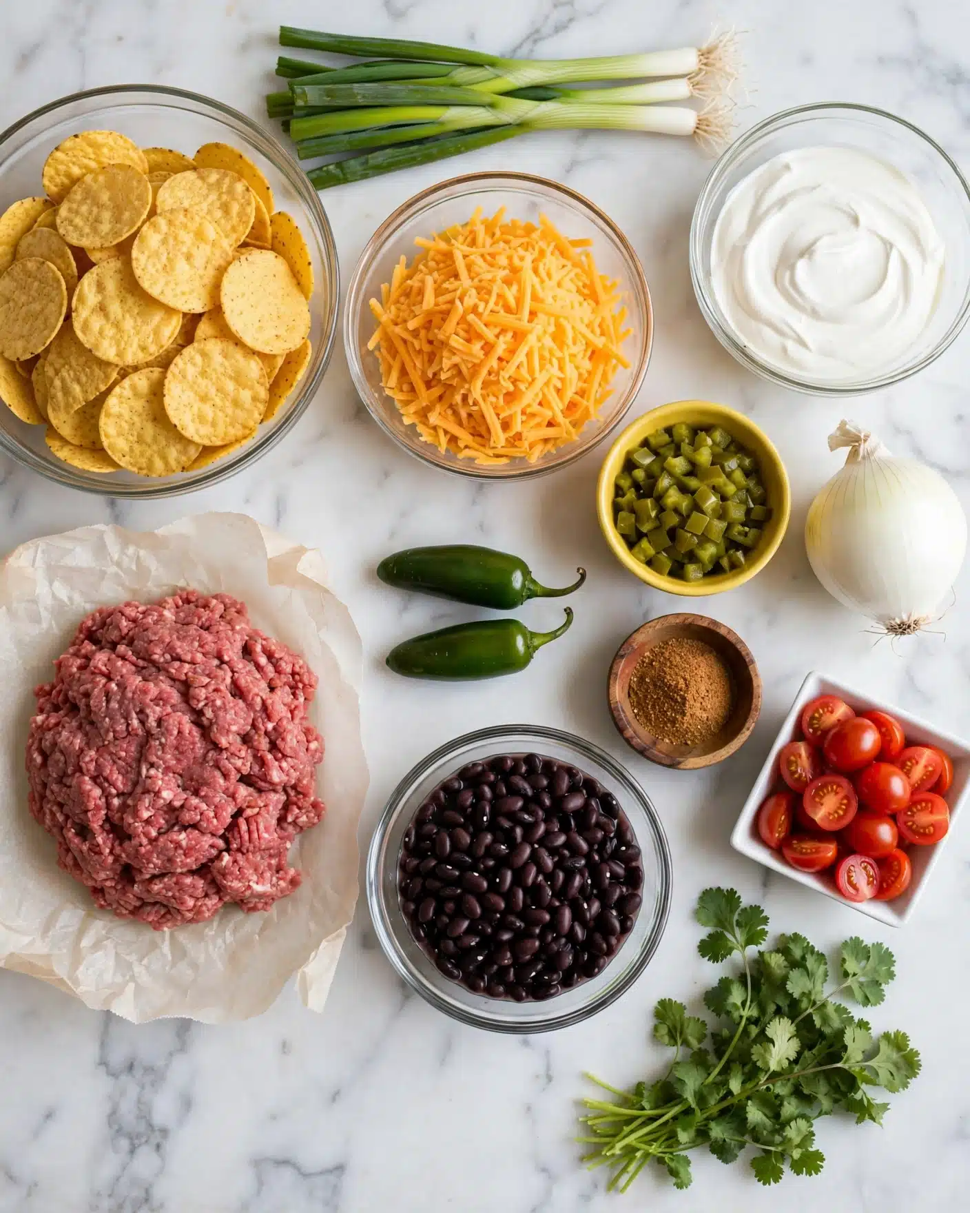 Best Nachos Supreme Recipe 4 The image shows separate ingredients arranged on a white marbled surface for making a layered dish. There is a clear bowl of yellow round chips on the top left and a mound of raw ground meat on white parchment below it. To the right, a clear bowl holds shredded orange cheese, while a small yellow bowl contains green diced pickled peppers. Two whole green jalapeños lay near the center. Fresh green onions are at the top, and cherry tomatoes fill a white square dish. A whole white onion sits near the right edge. A small wooden bowl of brownish-red spice mix is near the middle, and a glass bowl holds chunky black beans. Fresh cilantro with stems is at the bottom right, and a clear bowl of white sour cream is near the top right. No prepared layers are visible, just the ingredients spread out for assembly. Photo taken with an iphone --ar 4:5 --v 7